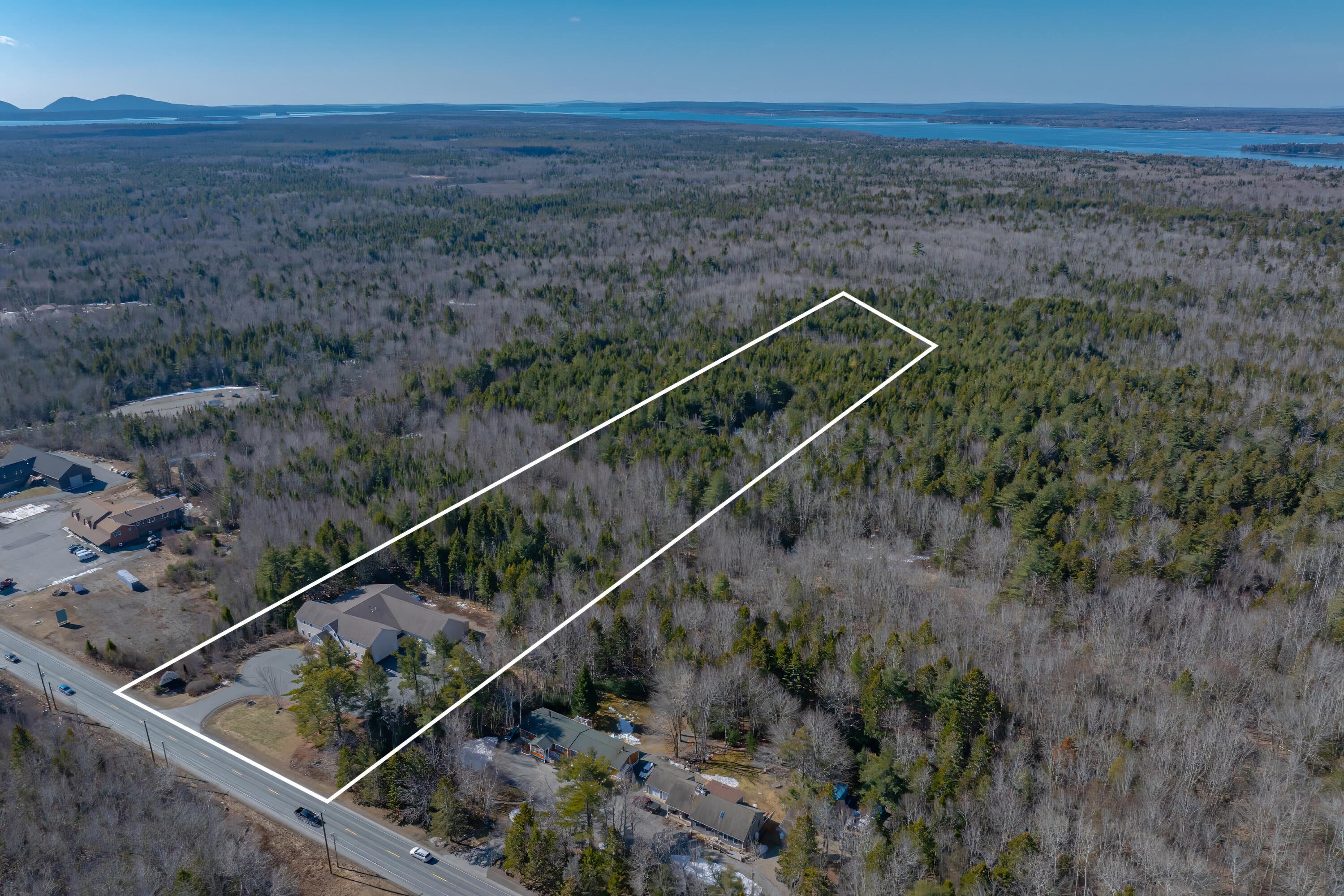 Aerial view of a wooded parcel outlined in white, with a building by the road and water in the distance.
