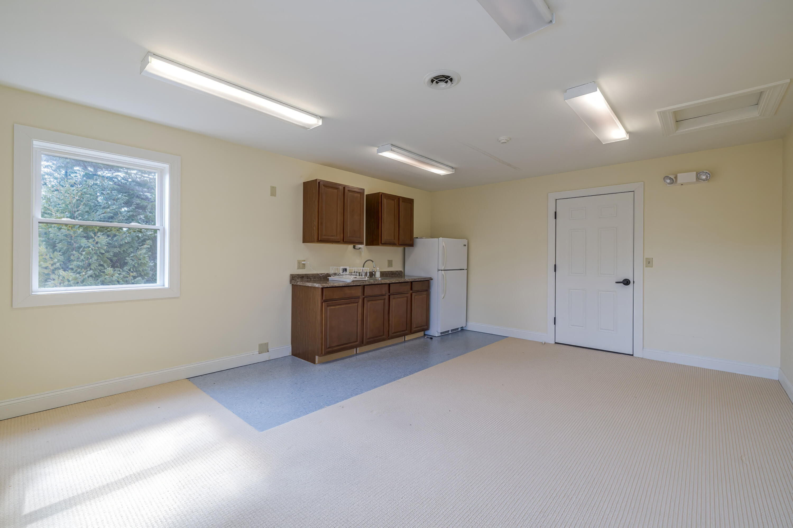 Vacant room with a wood-cabinet kitchenette, refrigerator, and large window overlooking trees.