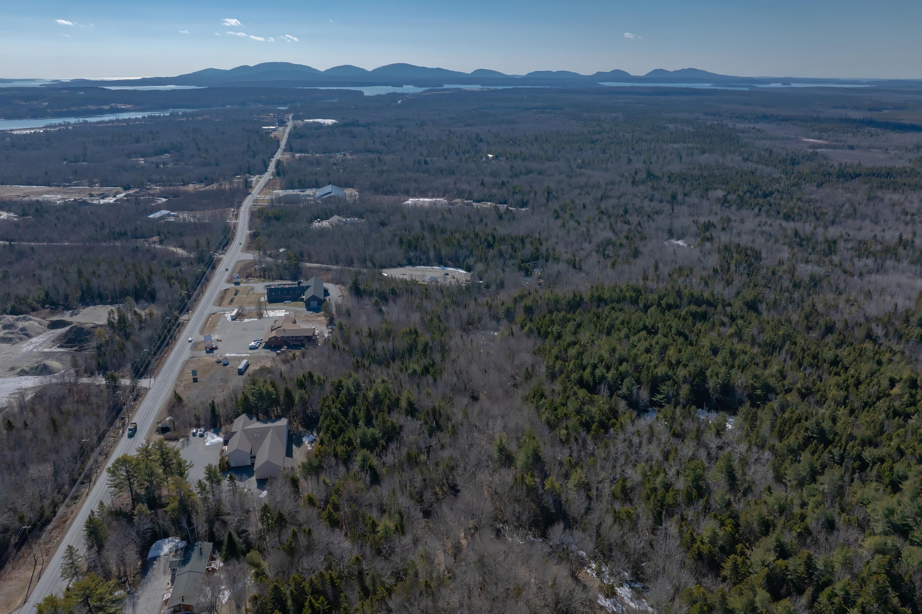 Aerial view of wooded land and buildings along a road, with distant water and hills.