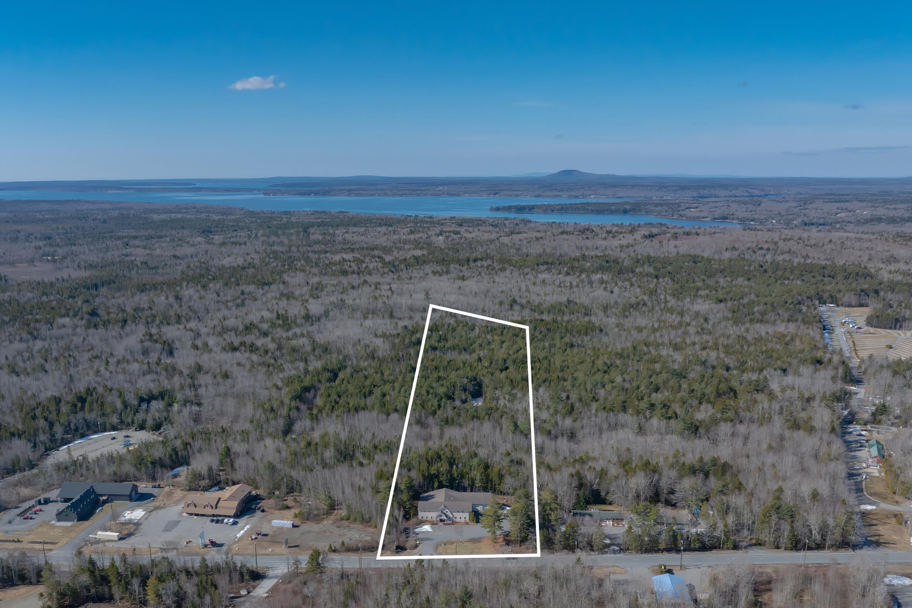 Aerial view of a wooded parcel outlined in white, with a building, road frontage, and distant water.