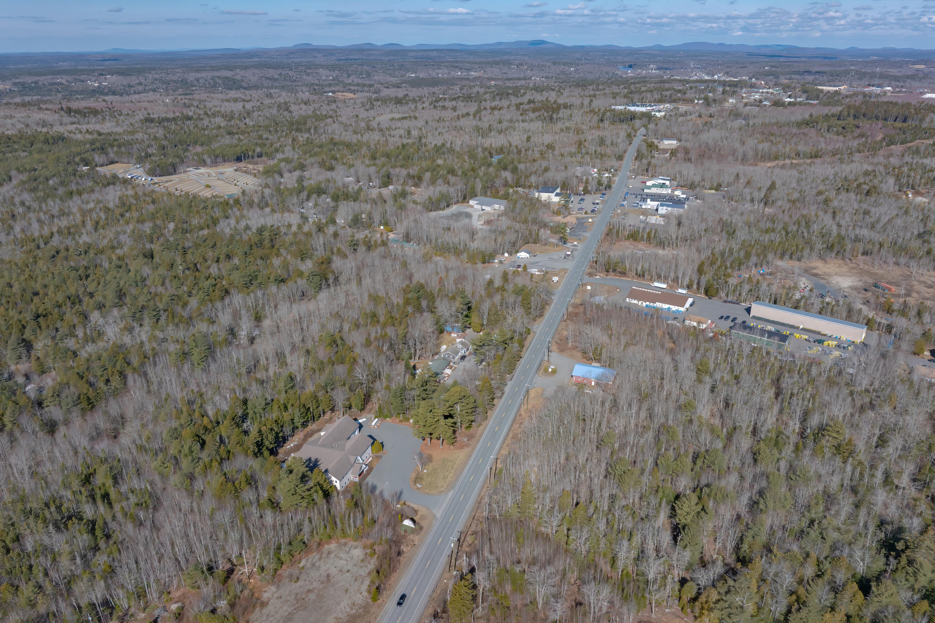 Aerial view of a wooded property beside Bar Harbor Road, with commercial buildings and distant hills.