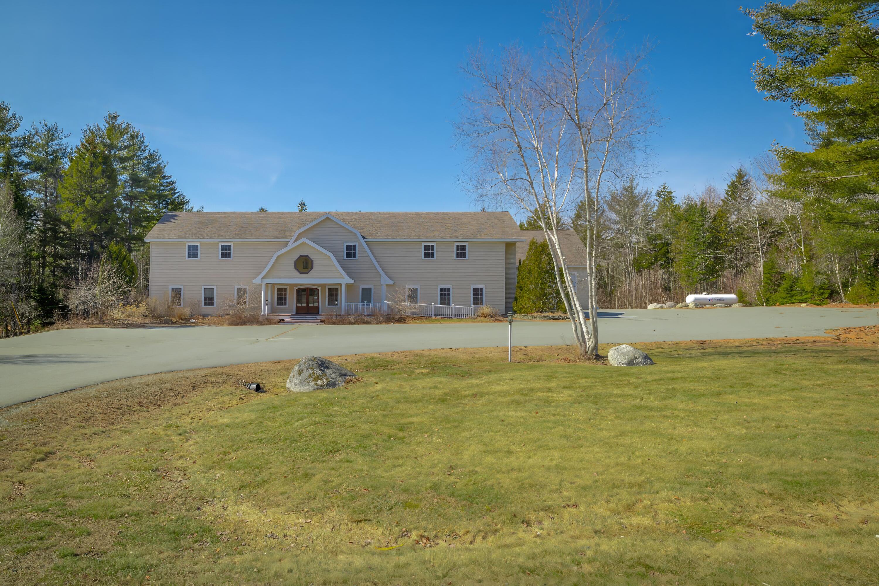 Large beige two-story building with a peaked entrance, broad driveway, and wooded surroundings.