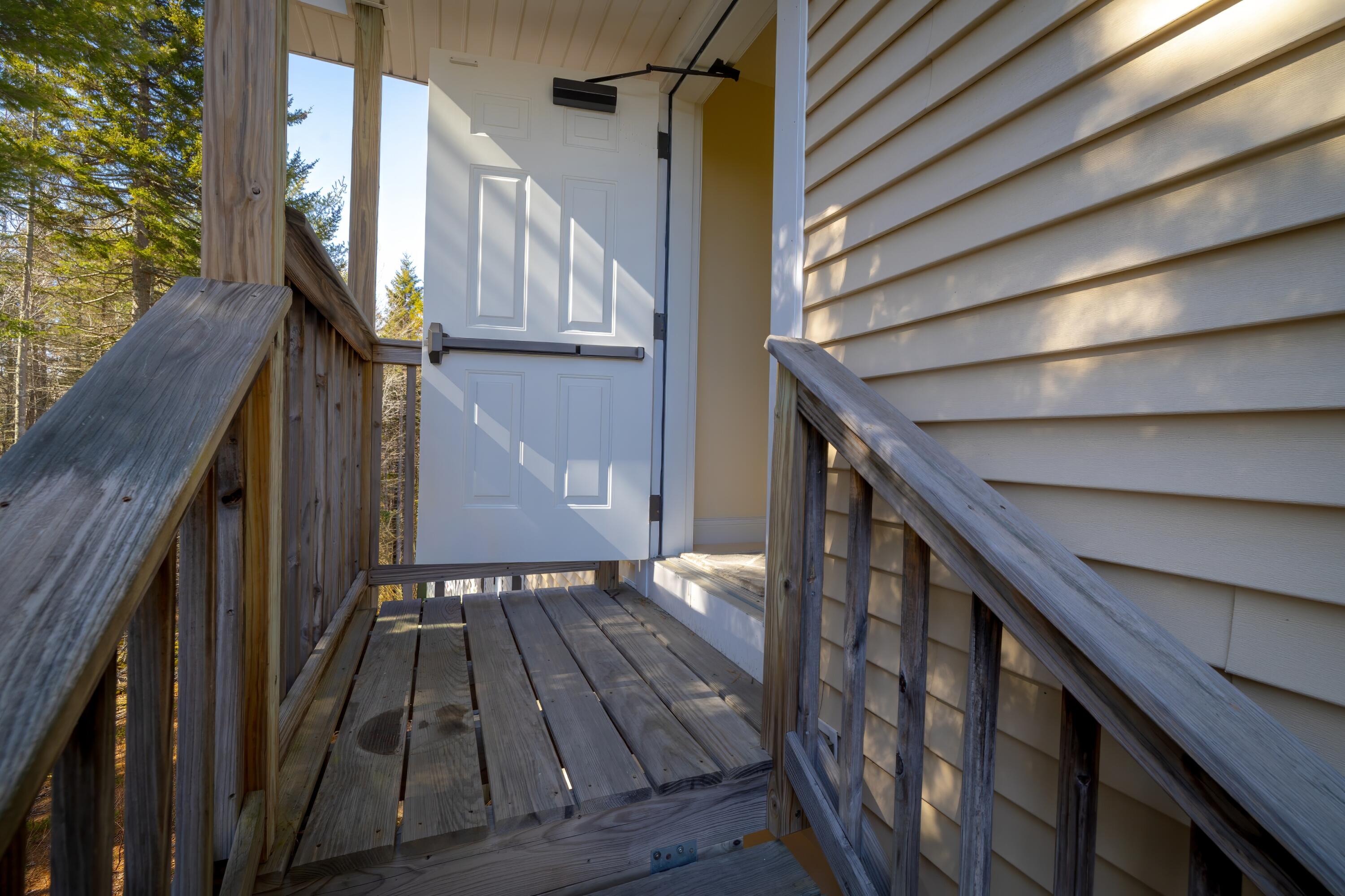 Home entrance with an open white door from a weathered wooden porch surrounded by trees.