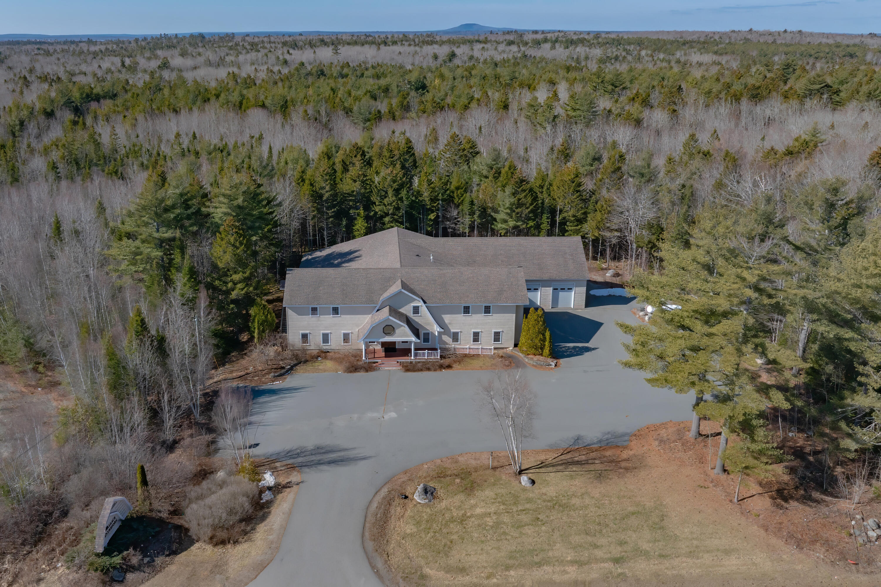 Aerial view of a large gray building and paved drive surrounded by woodland, with a distant mountain ridge.