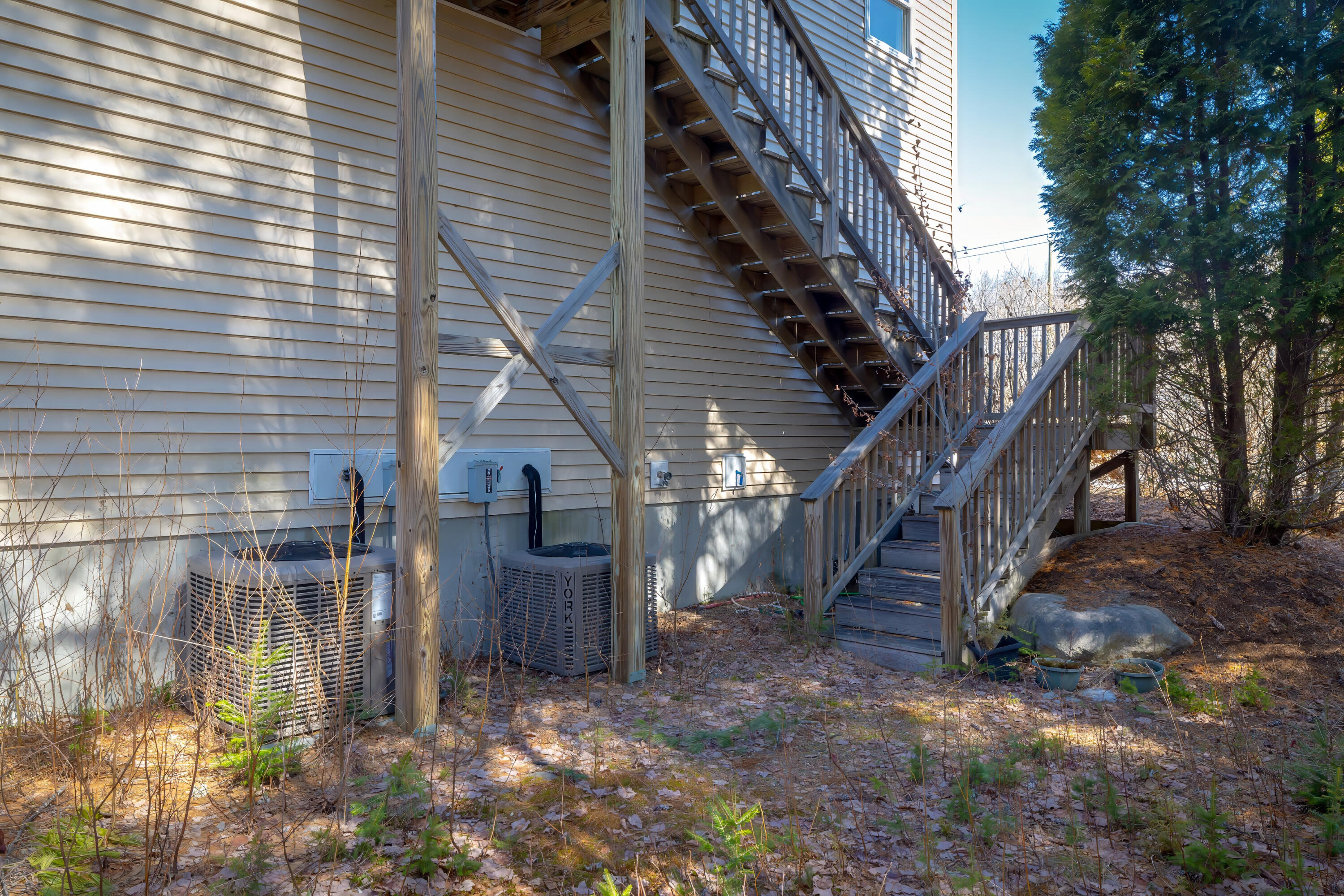 House exterior with wooden stairs and deck, two air-conditioning units, and leaf-covered ground.