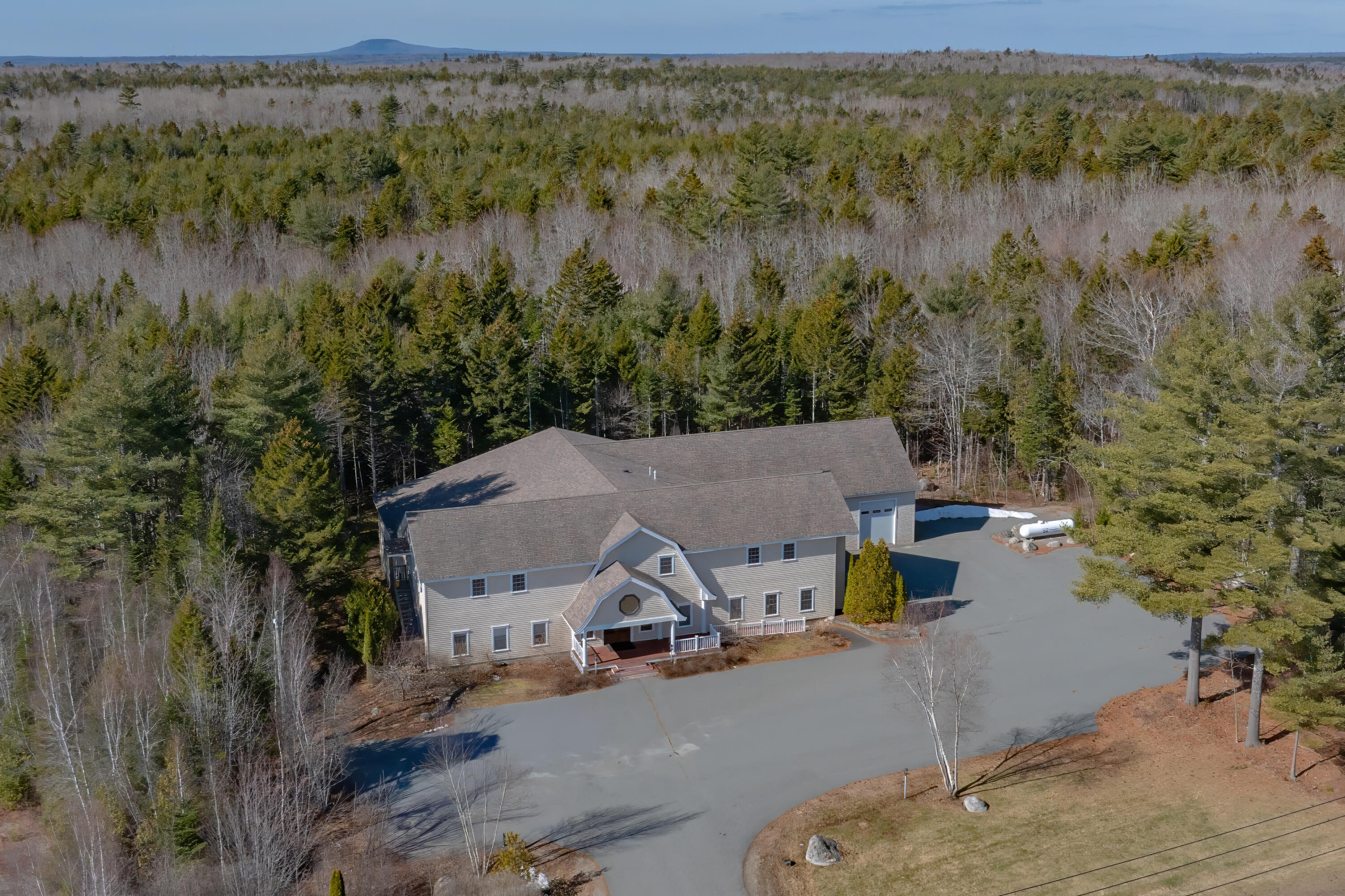 Large gray building and paved driveway surrounded by woodland, with a mountain visible in the distance.