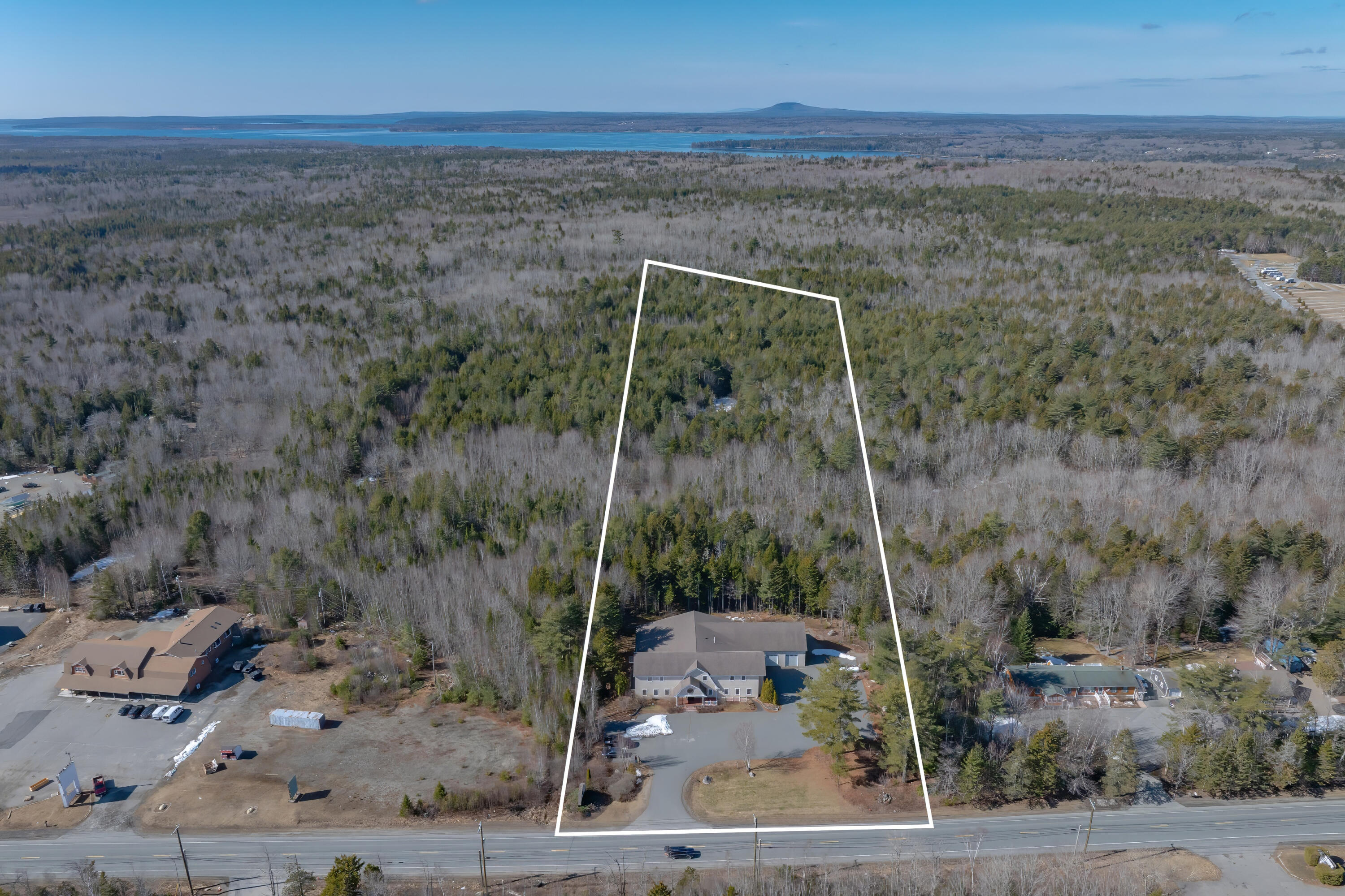Aerial view of a wooded parcel outlined in white, with a building near the road and distant water.