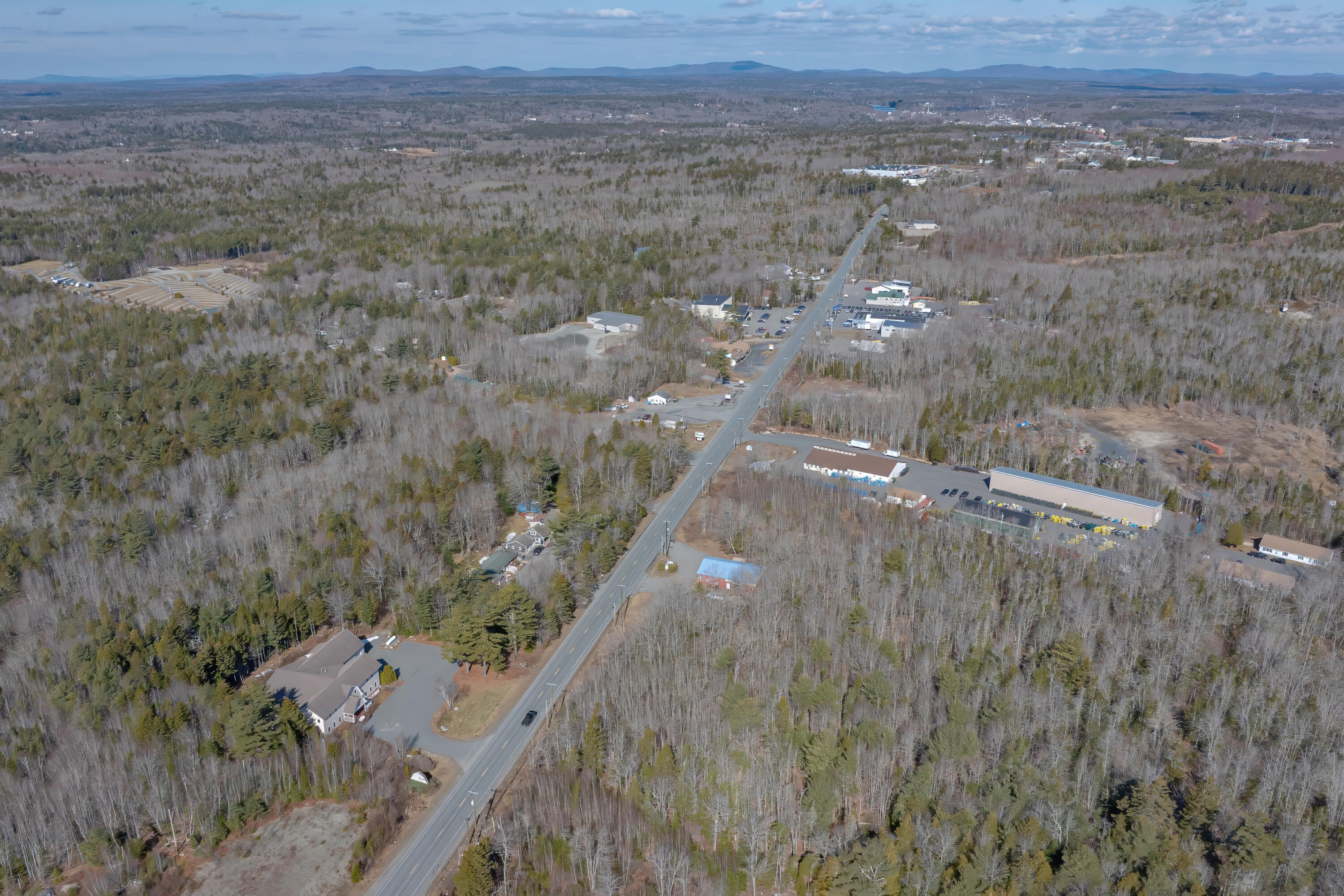 Aerial view of a road through leafless woods, with nearby homes, commercial buildings, and distant hills.