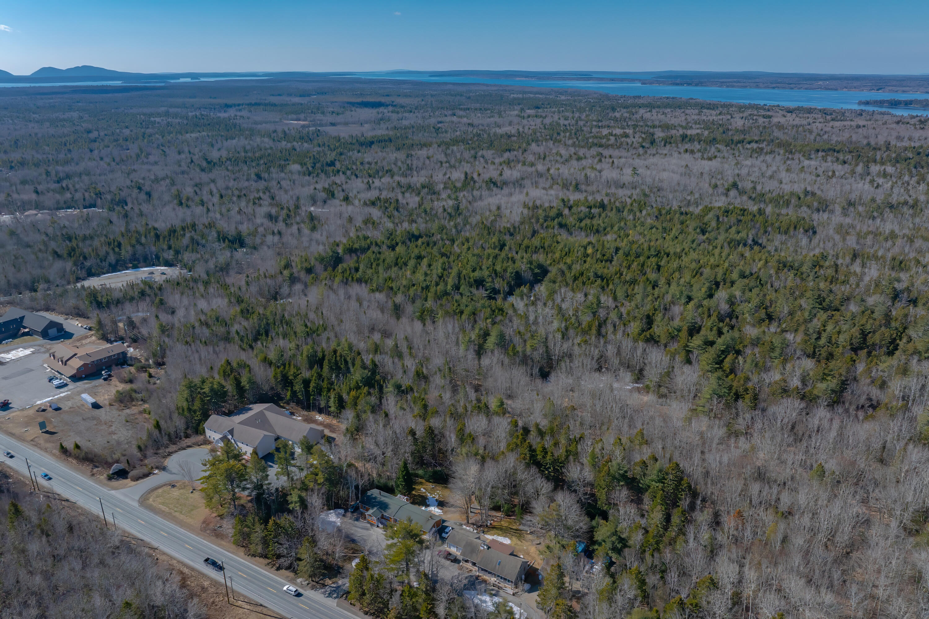 Aerial view of wooded property beside a road, with buildings, distant water, and low hills.