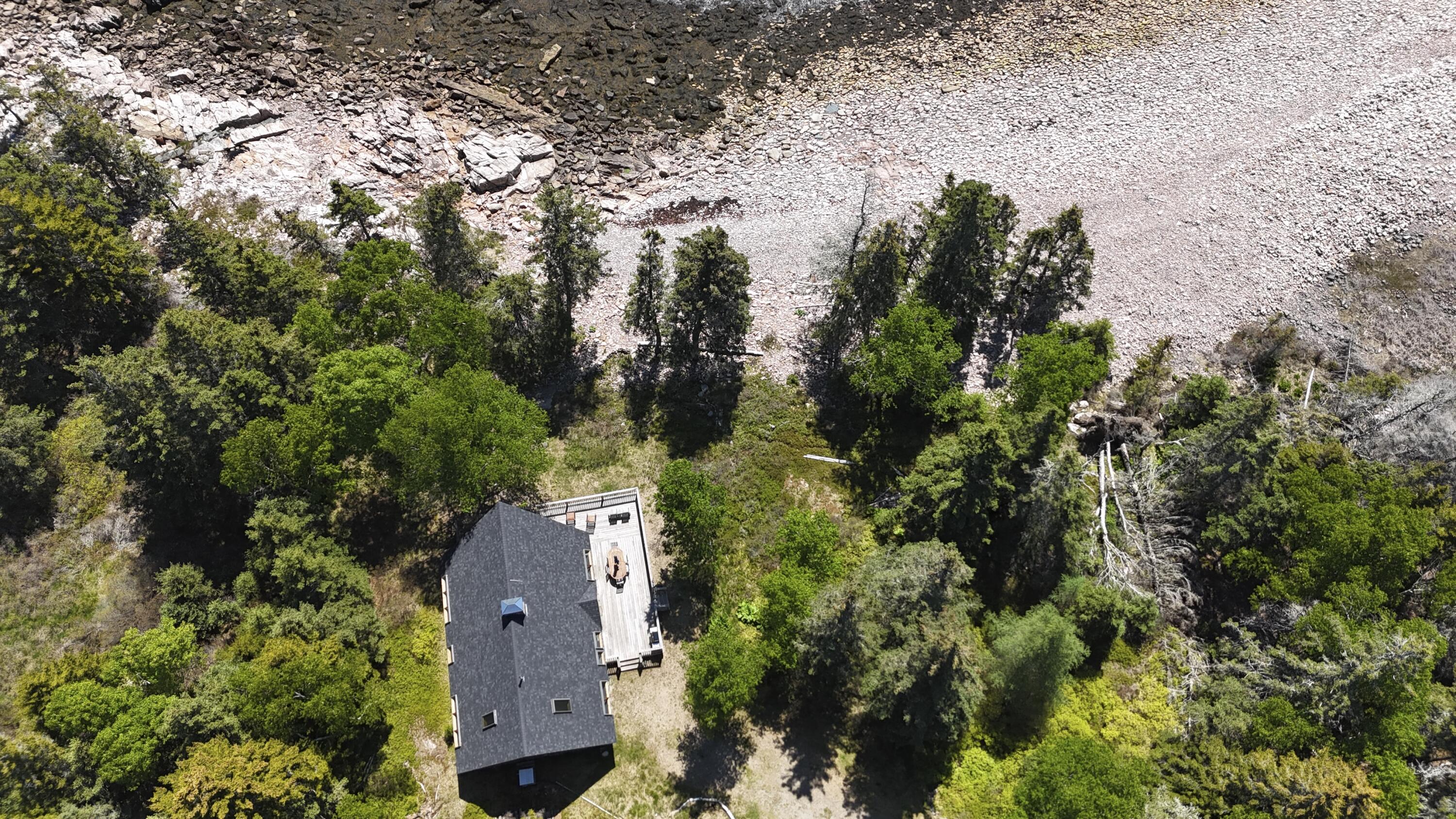 Aerial view of a wooded house beside a rocky shoreline, with a deck and outdoor furniture.