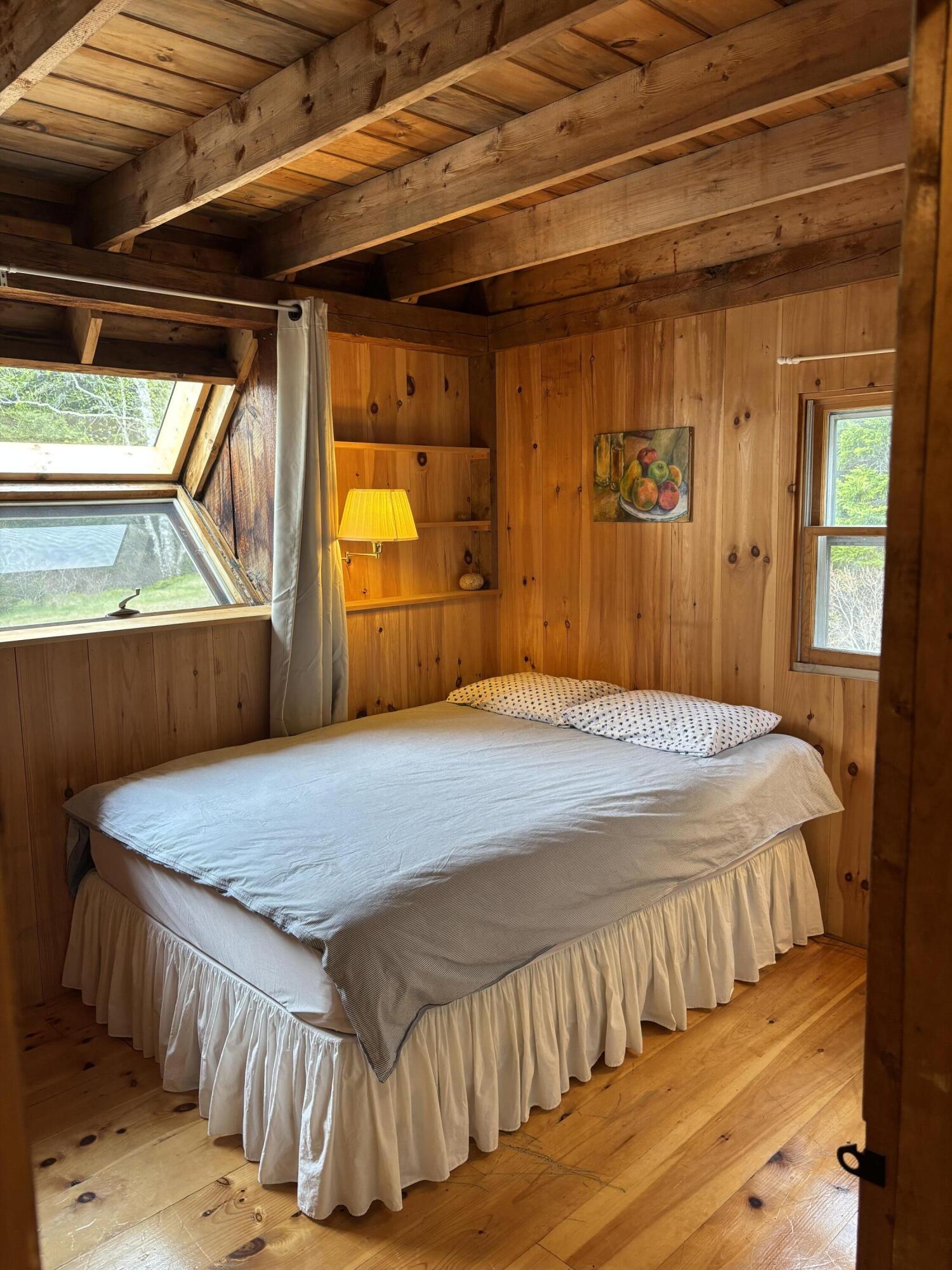 Pine-paneled bedroom with a double bed beneath exposed timber ceiling beams and windows overlooking greenery.
