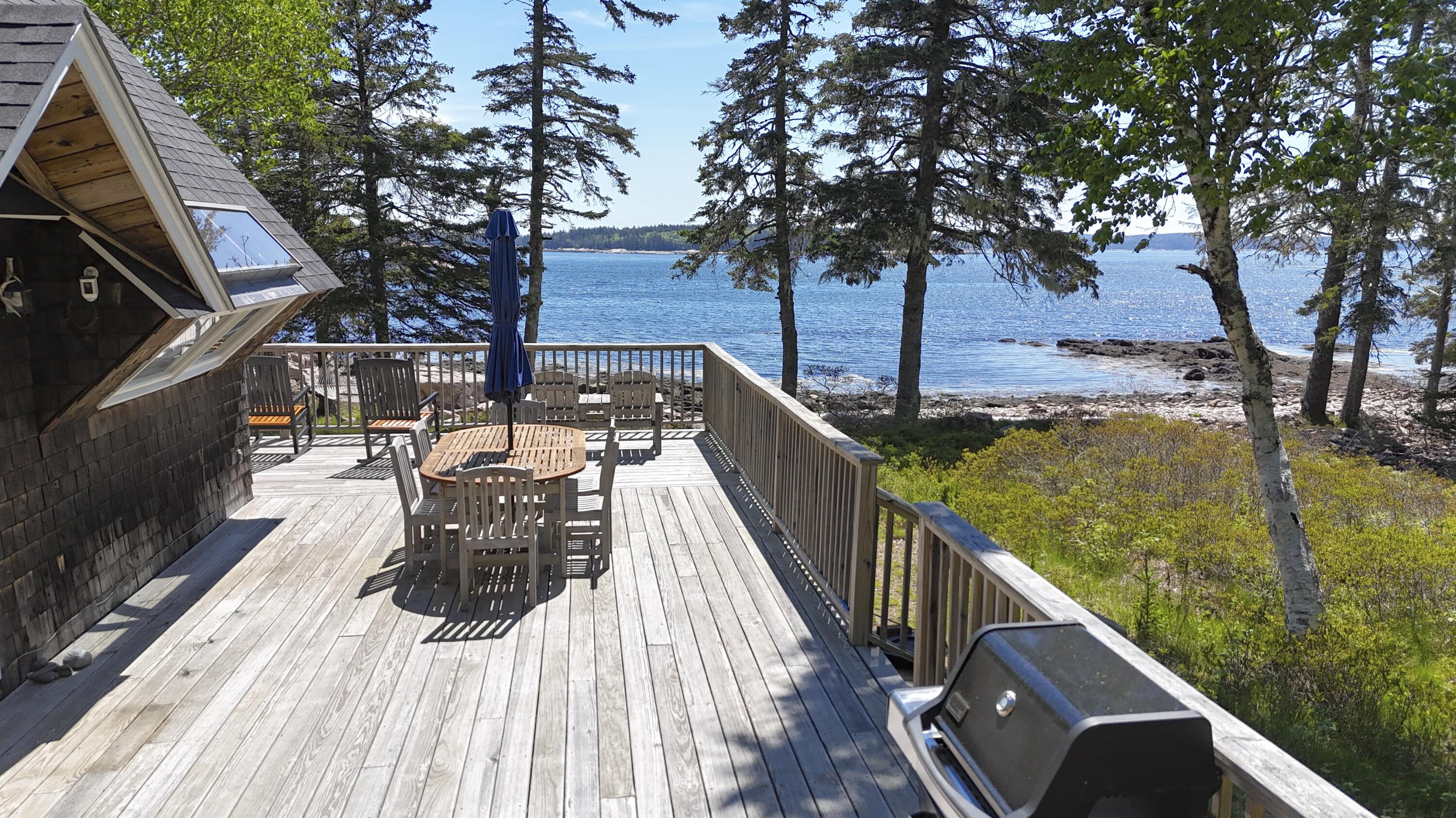 Furnished wooden deck with dining set and grill overlooking a rocky shoreline and blue water