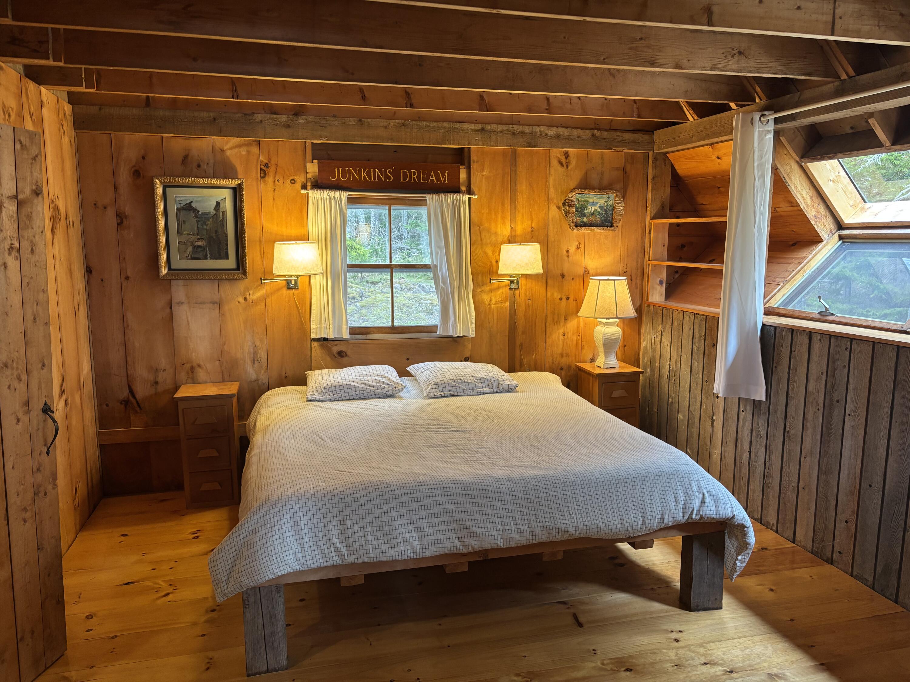 Bedroom with a large bed, warm wood paneling, exposed beams, and a slanted window overlooking trees.