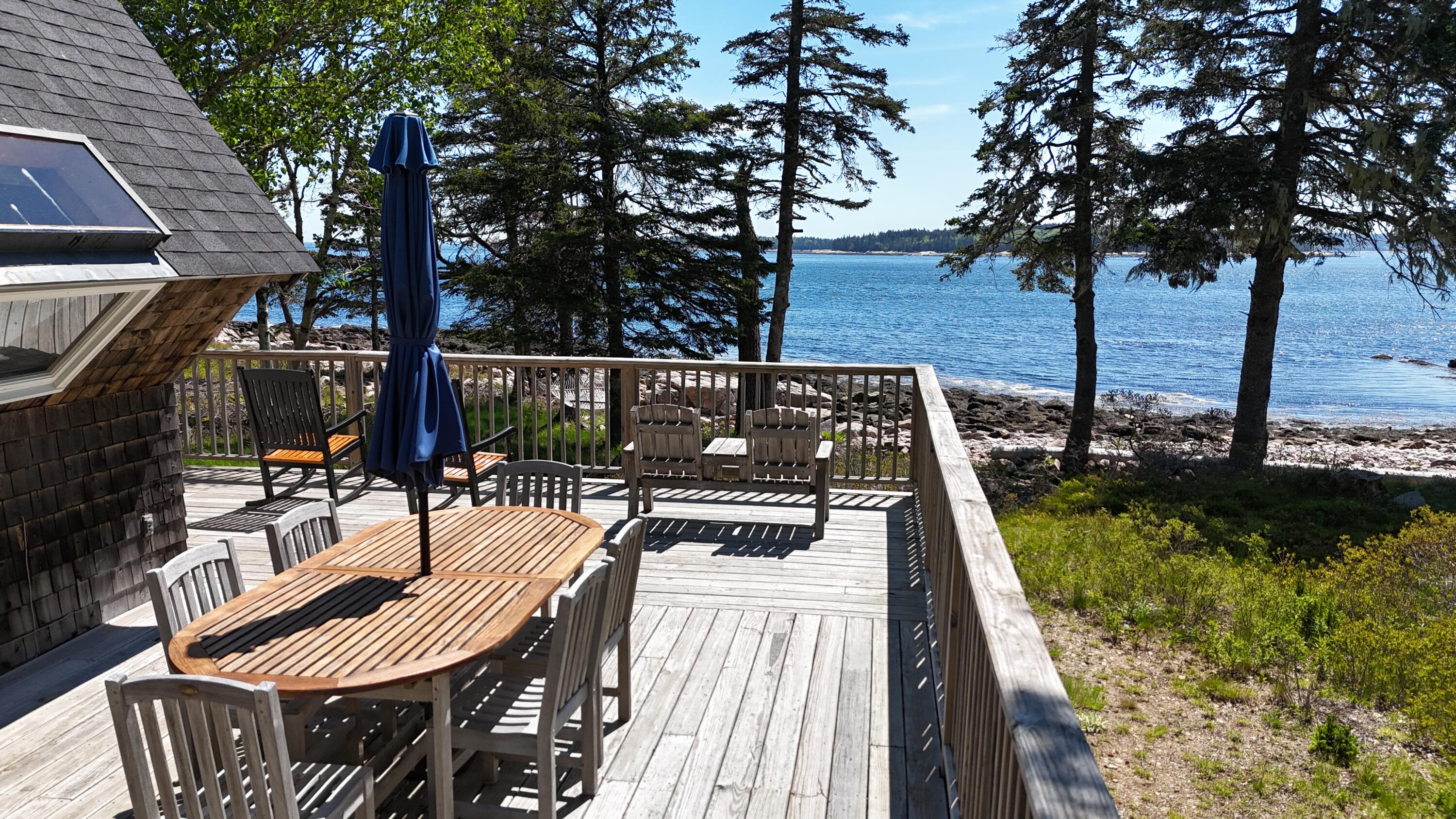Furnished waterfront deck with outdoor dining table, evergreen trees, and rocky shoreline.