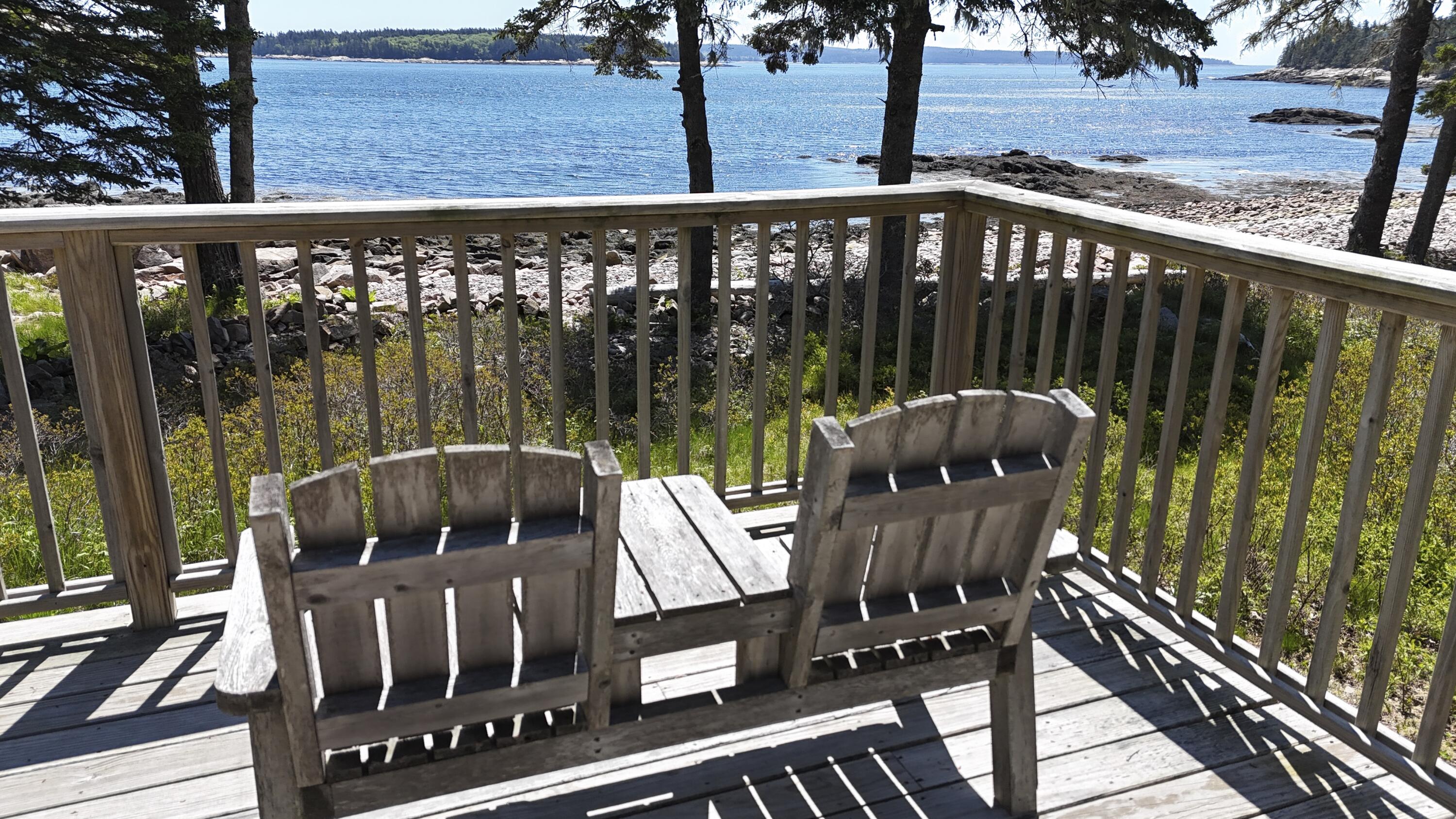 Oceanfront deck with weathered wooden chairs overlooking rocky shore and blue water.