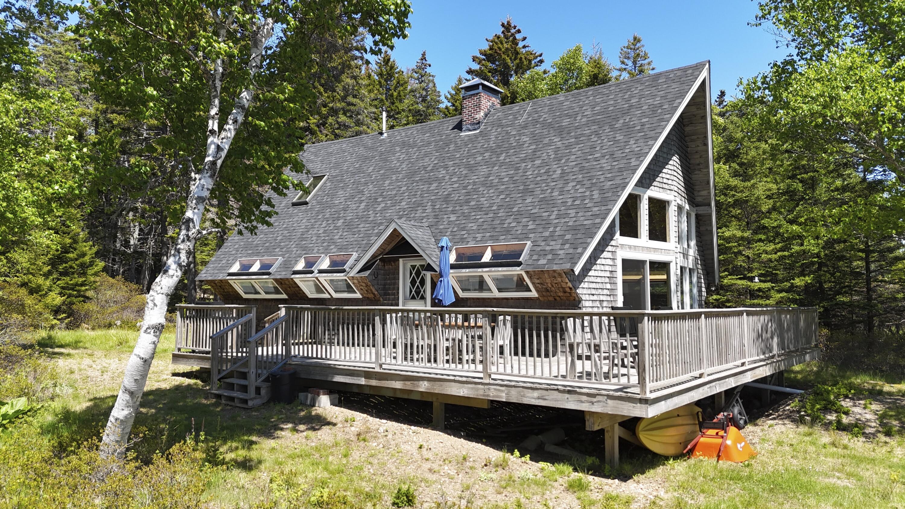 Woodland house with a steep gray roof, broad raised deck, patio furniture, and kayaks stored underneath.