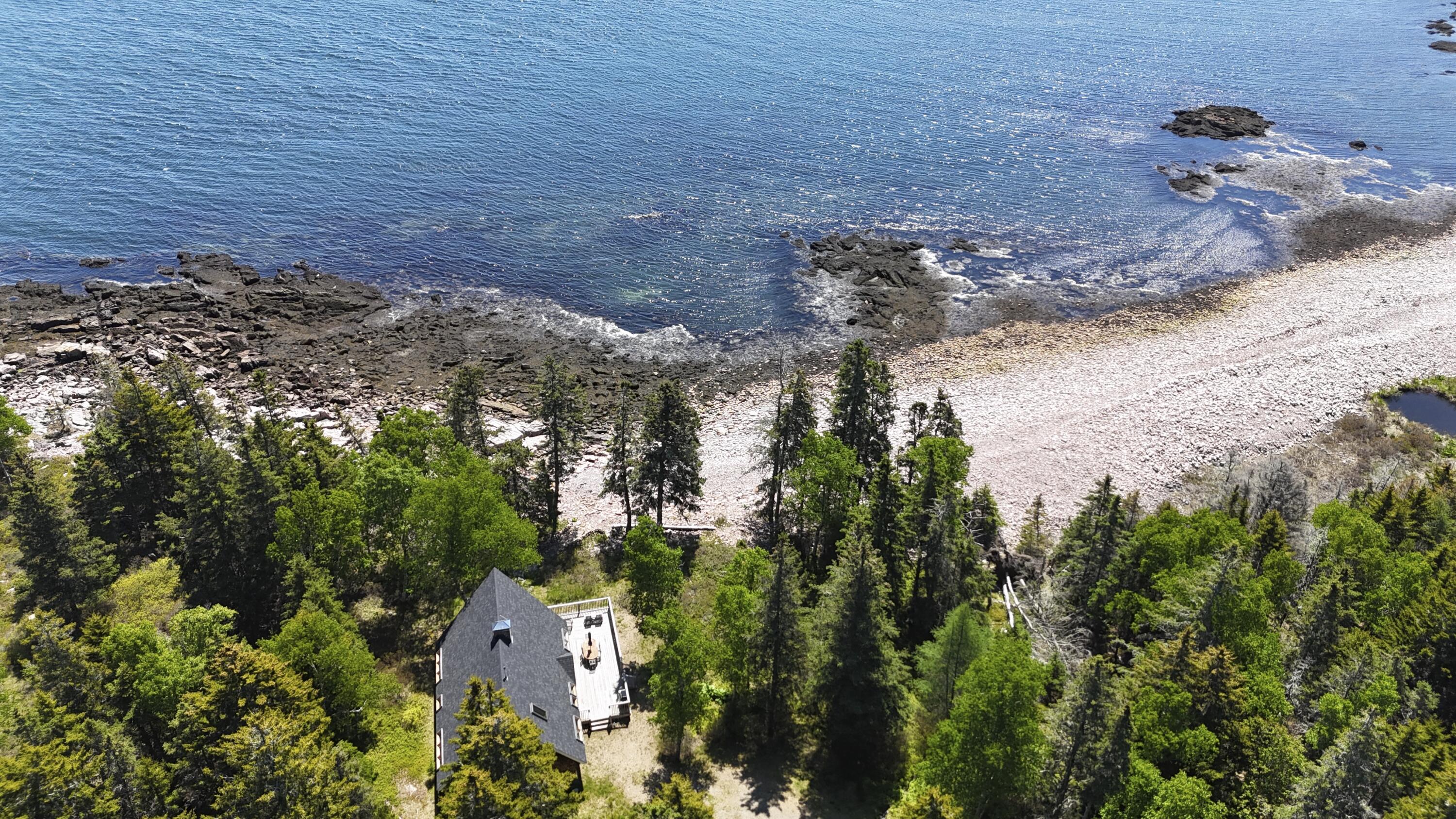Aerial view of a wooded oceanfront home beside a rocky beach and rugged shoreline.