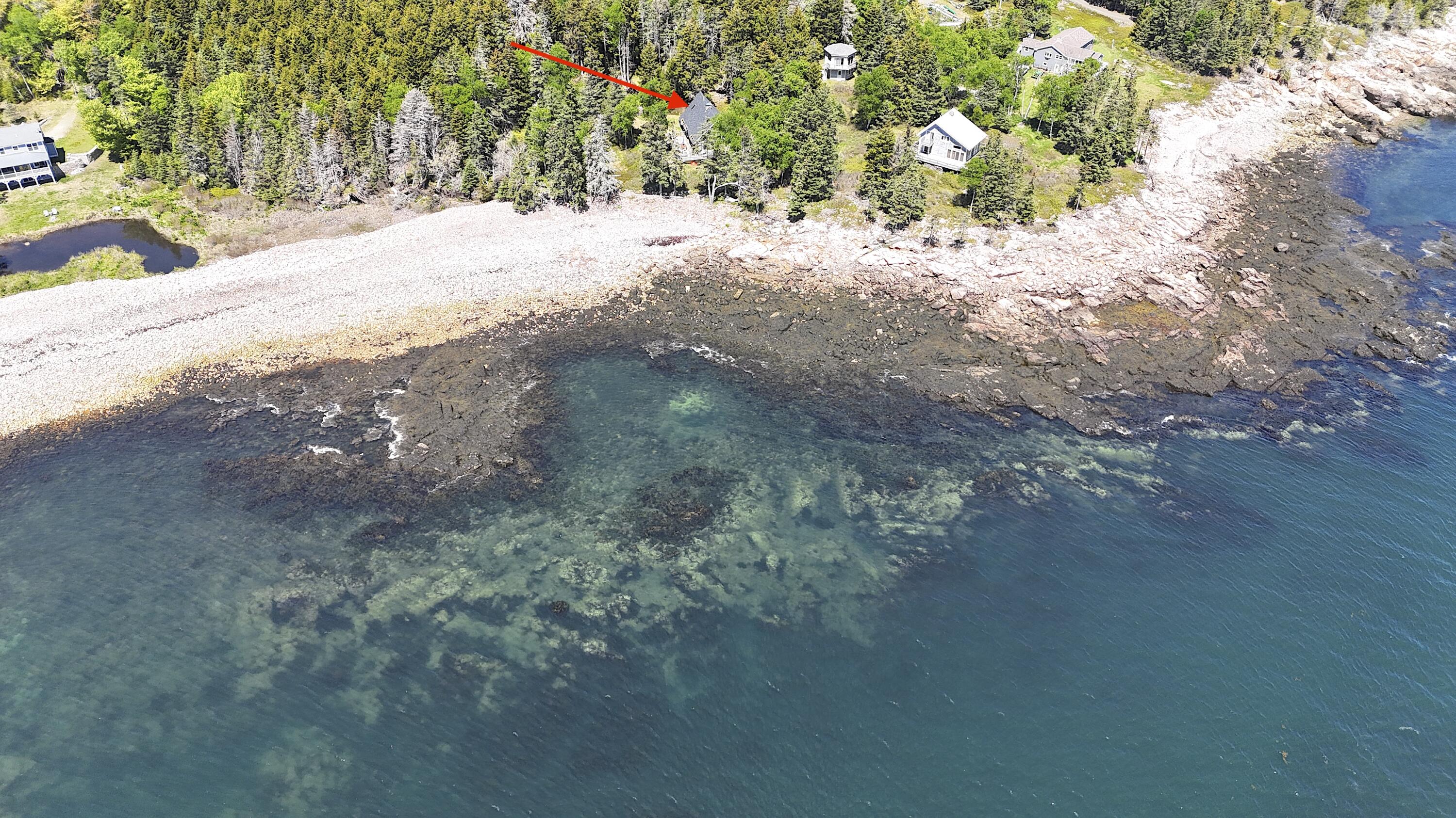 Aerial view of a wooded coastal property, marked by a red arrow, beside a rocky shore and clear water.