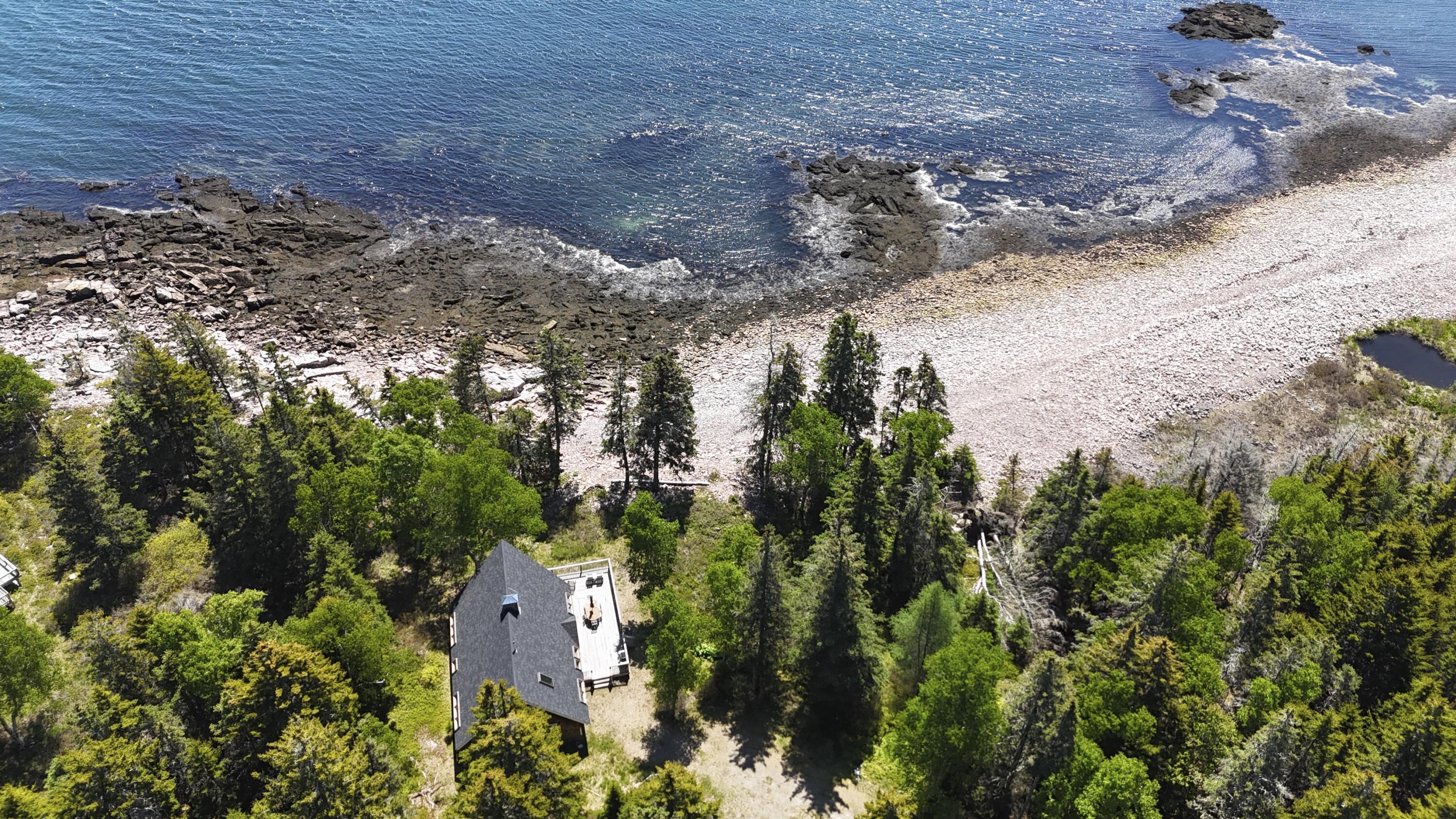 Aerial view of a wooded coastal home beside a rocky shore and blue water.