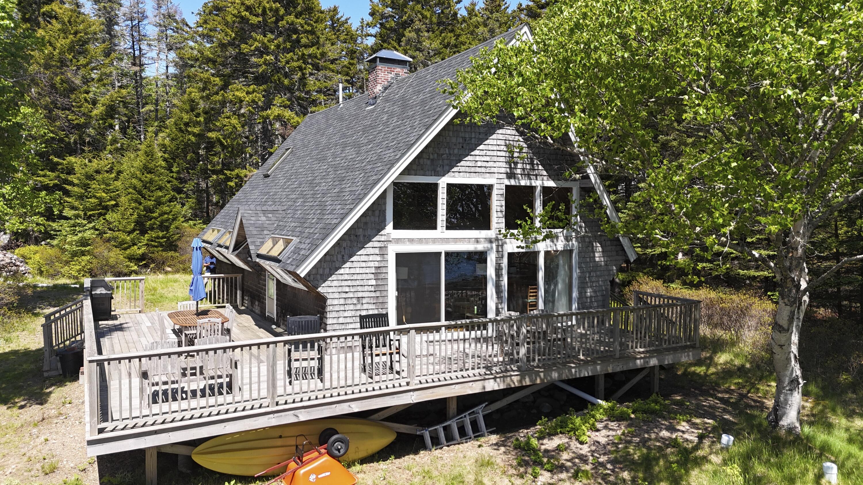 Gray-shingled A-frame house with a wide furnished deck and kayaks stored underneath.