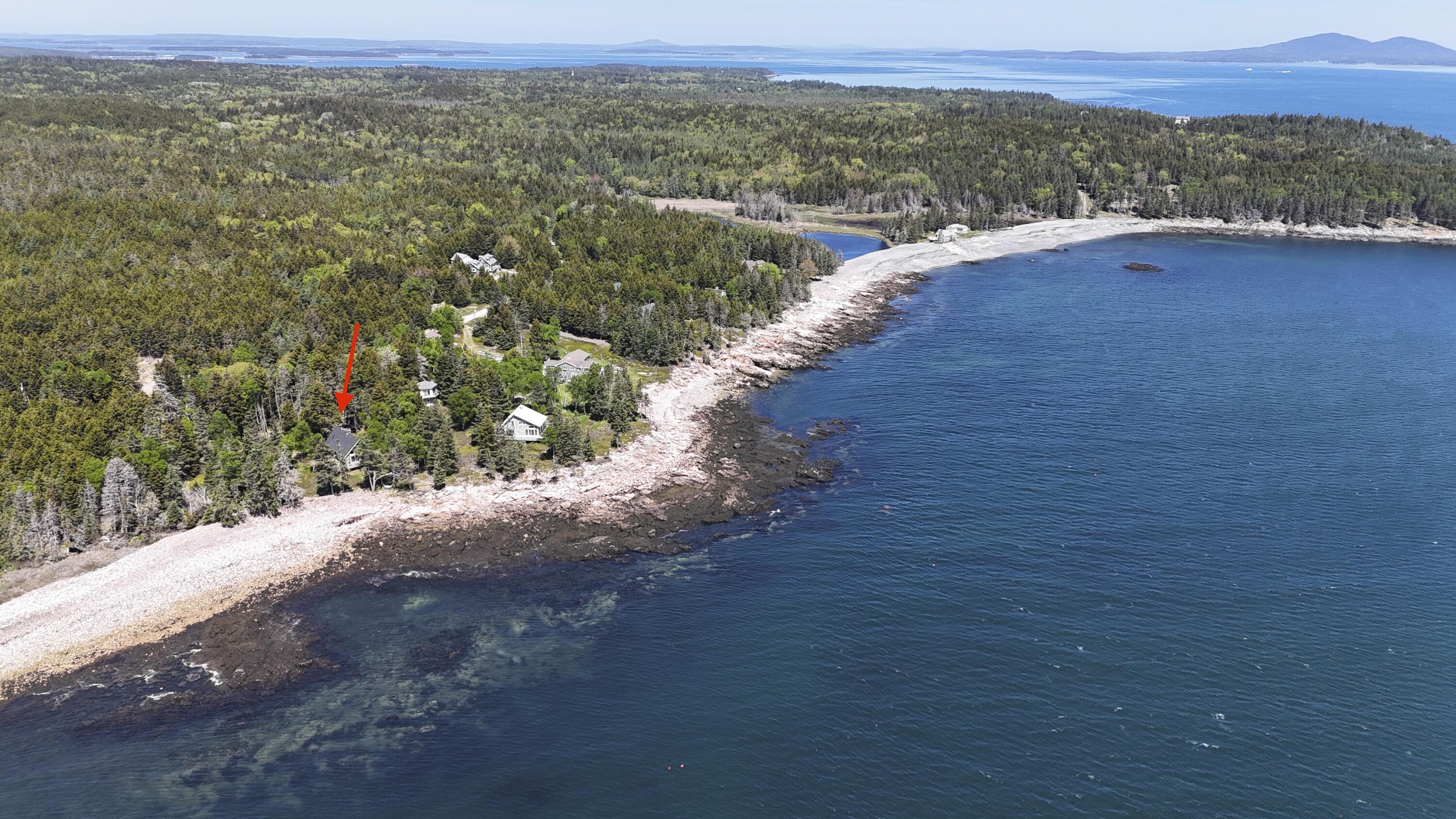Aerial view of a wooded coastal home marked by a red arrow beside rocky shore and open water.