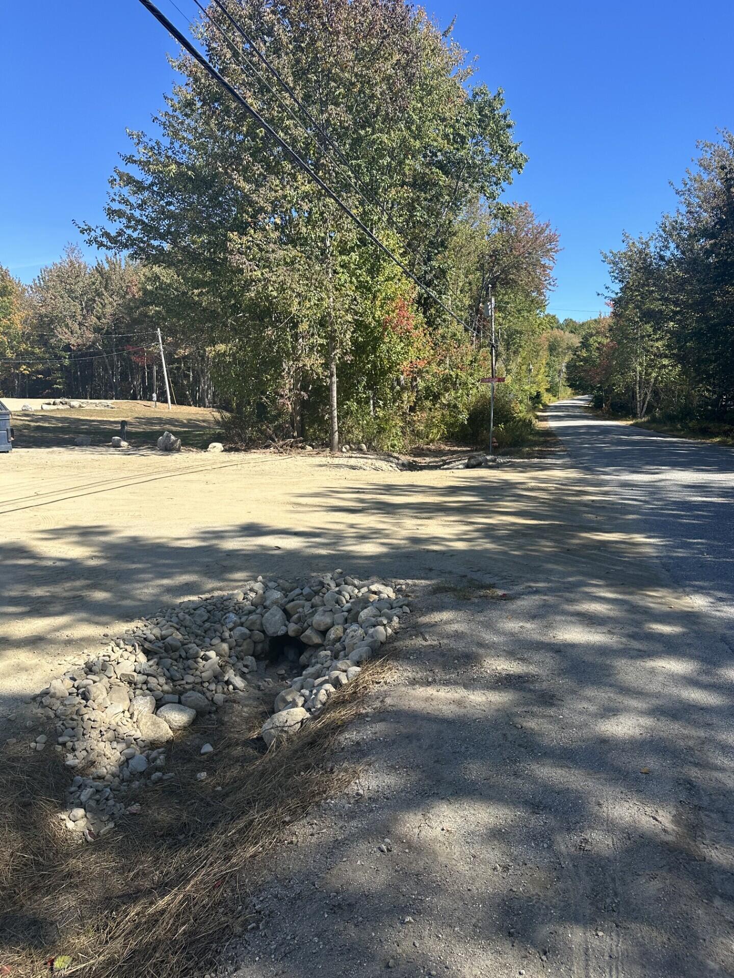 Gravel road beside a wooded vacant lot, with a stone-lined culvert in the foreground.