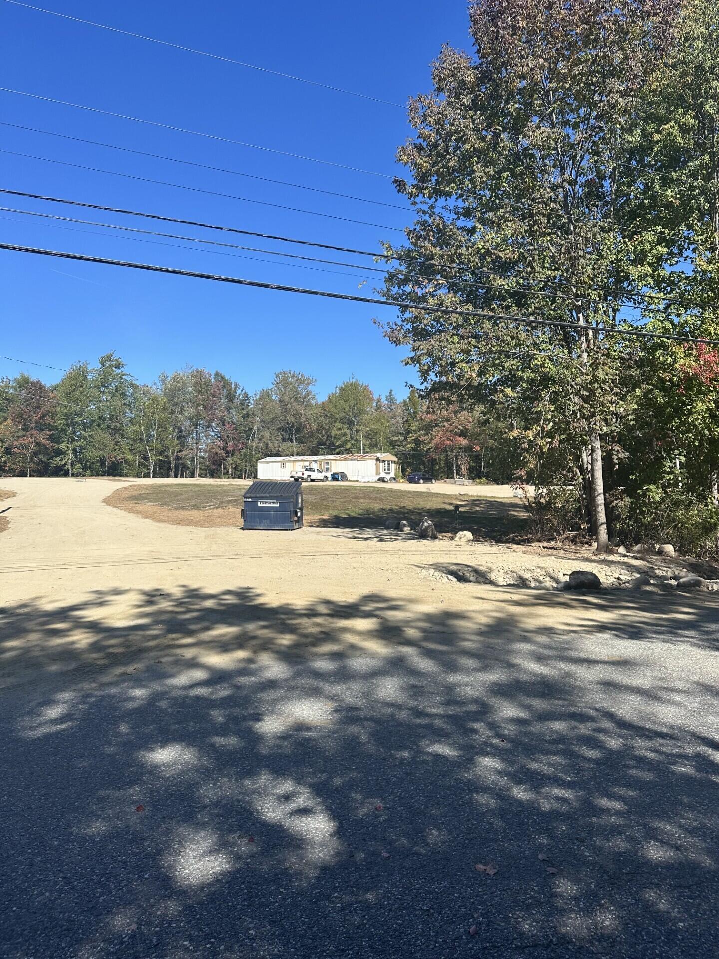 Graded property lot with a gravel drive, dumpster, and trees; a manufactured building stands in the distance.
