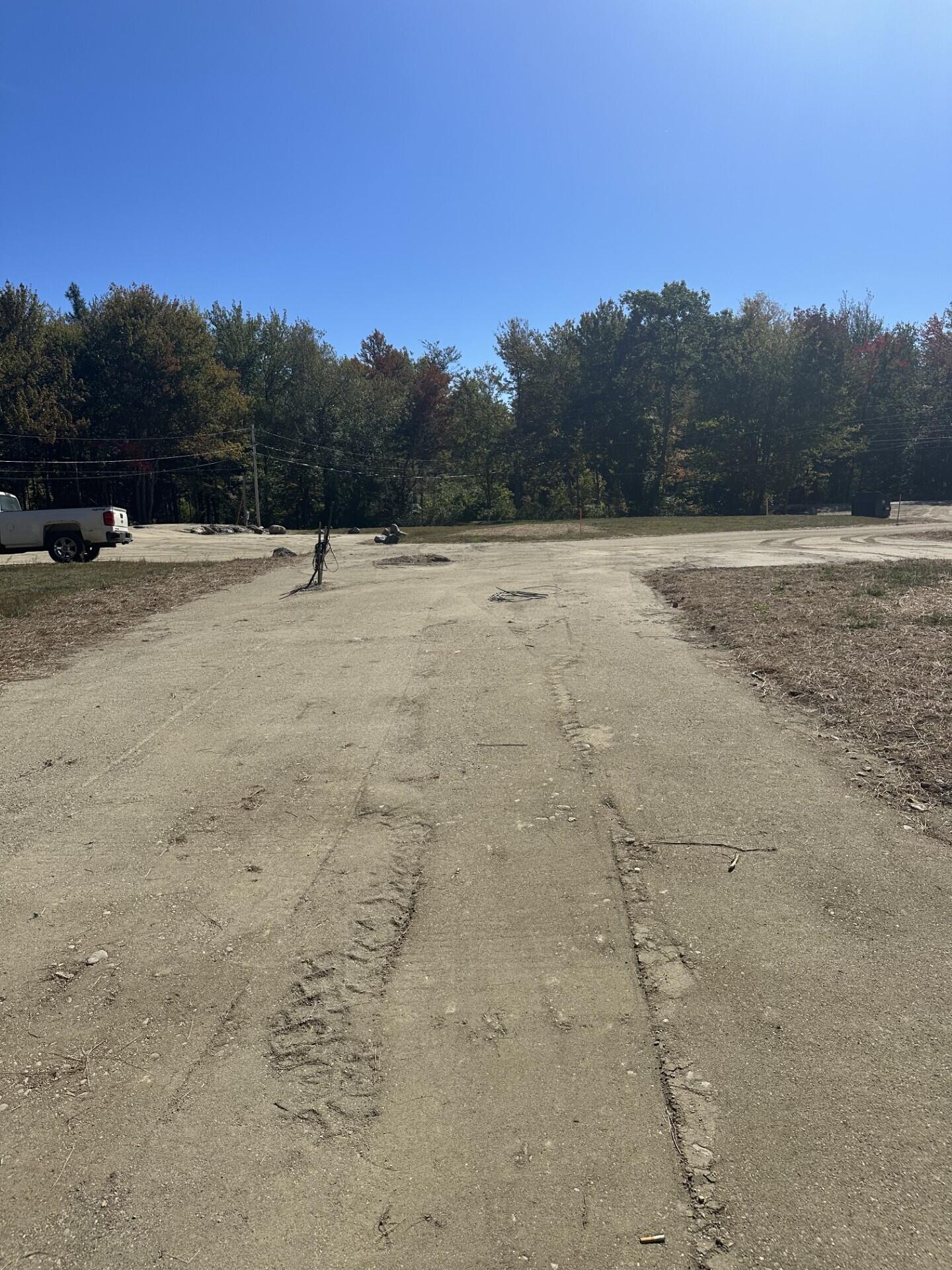 Cleared gravel lot bordered by trees, with utility wires and a white pickup at the left.