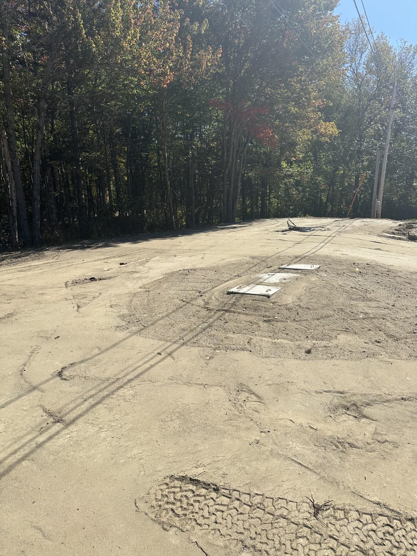 Cleared vacant lot bordered by autumn woods, with utility covers on the graded ground.