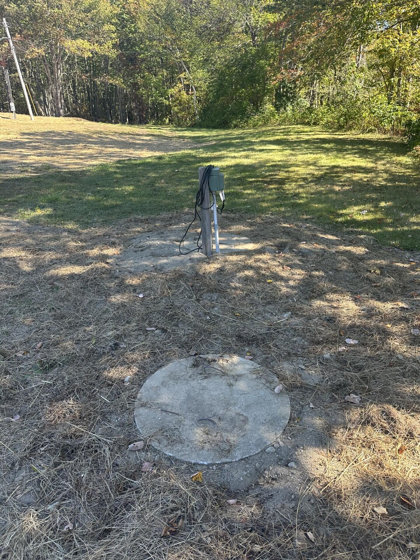 Cleared lot bordered by trees, with utility equipment and a round concrete cover.