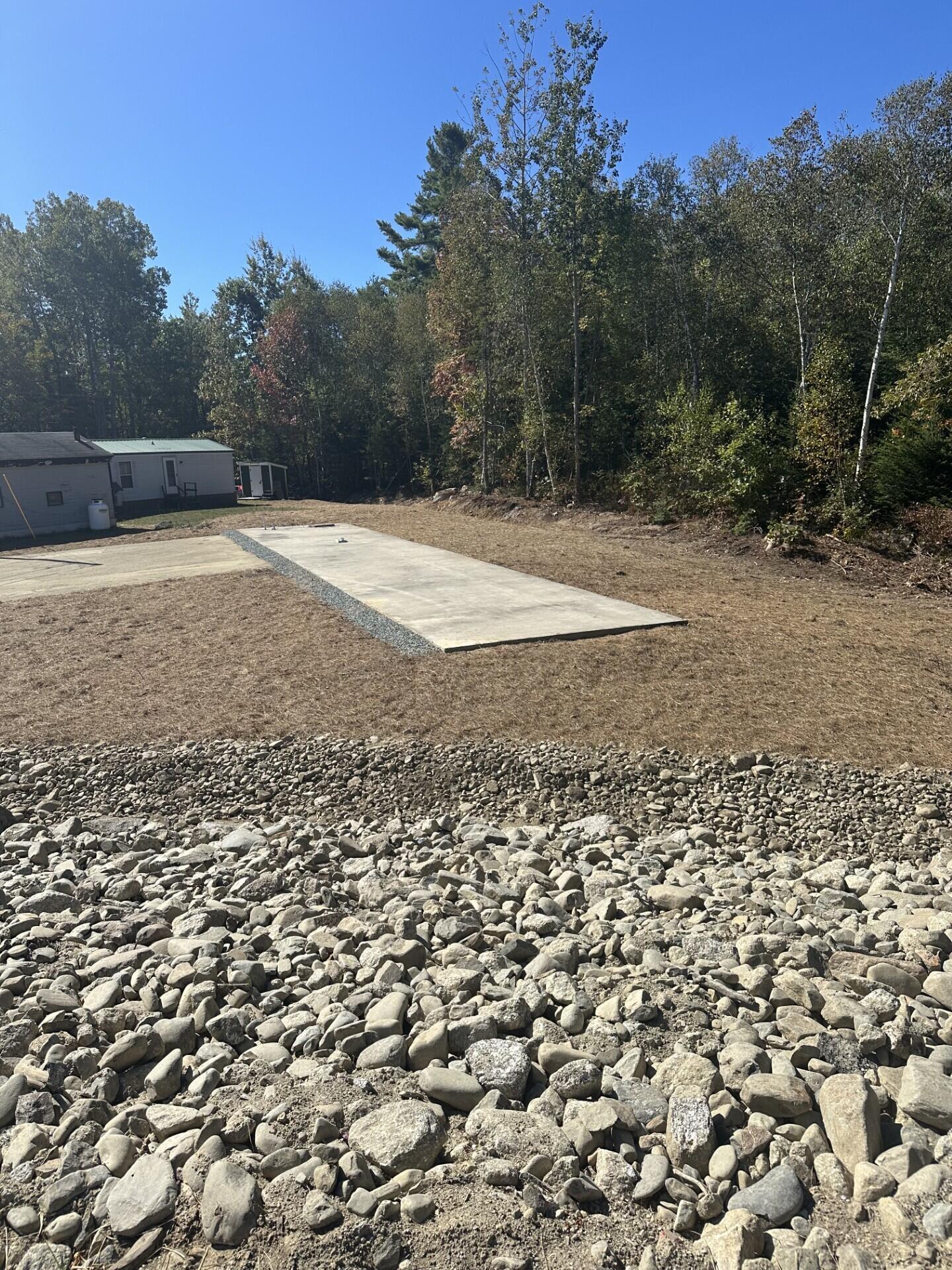 Cleared wooded lot with a rectangular concrete pad, rocky foreground, and a small manufactured home at left.