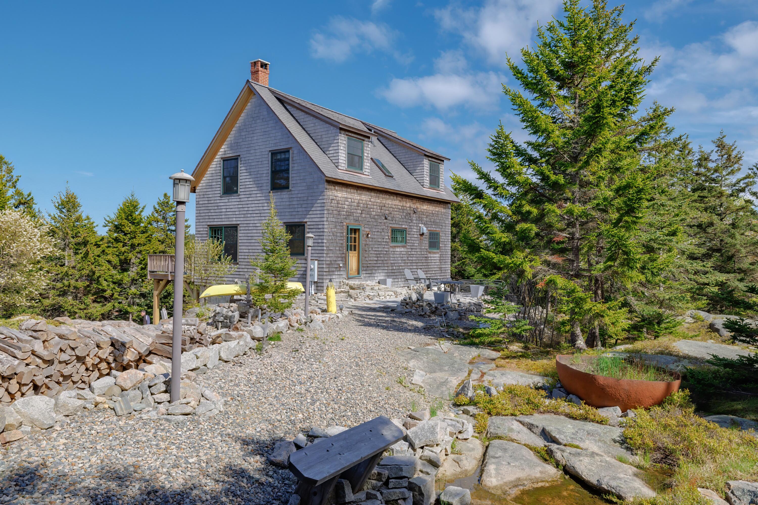 Gray shingle-sided house with a steep roof among evergreens, rocks, and gravel landscaping.