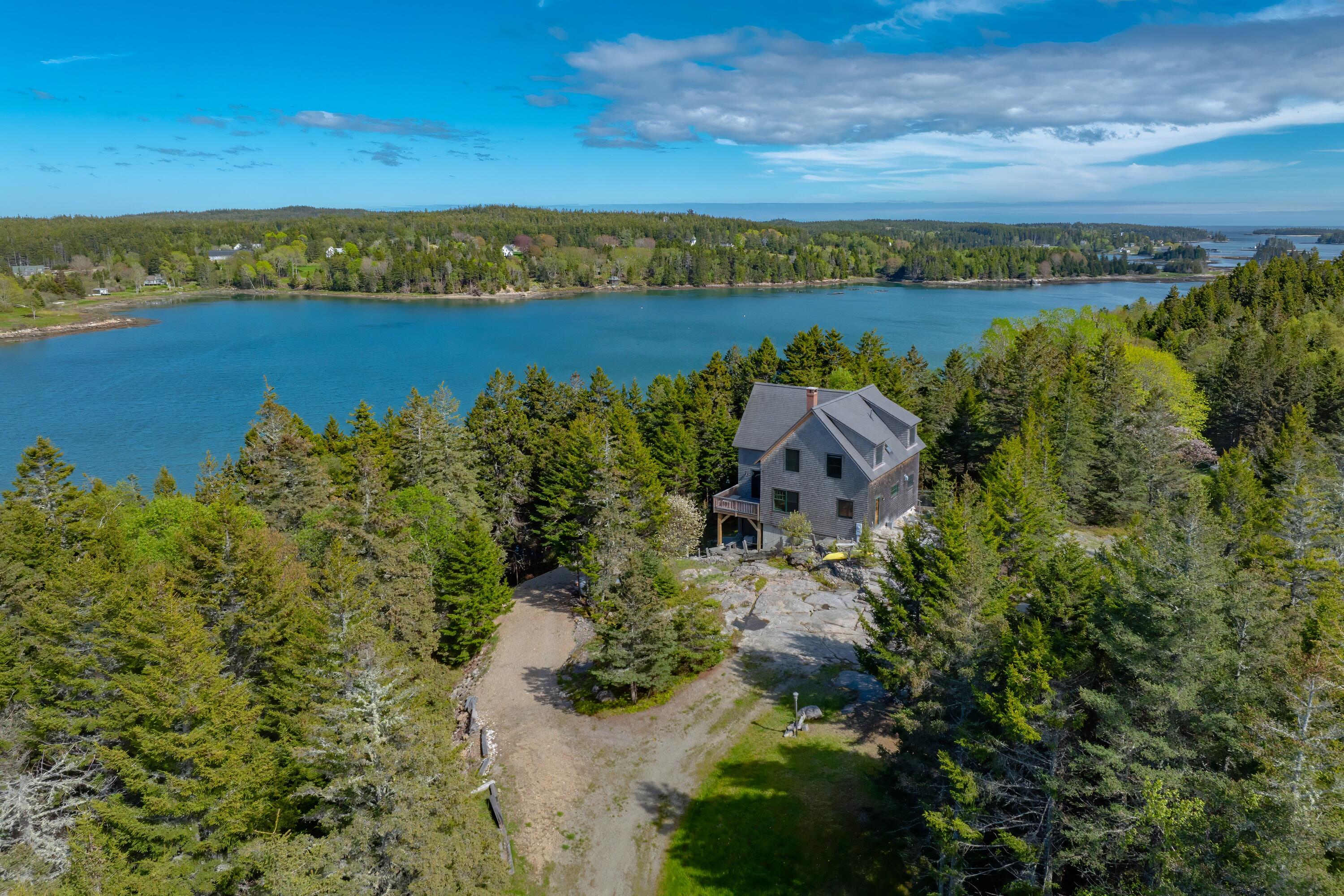 Aerial view of a coastal house among trees beside a blue inlet and rocky shoreline.