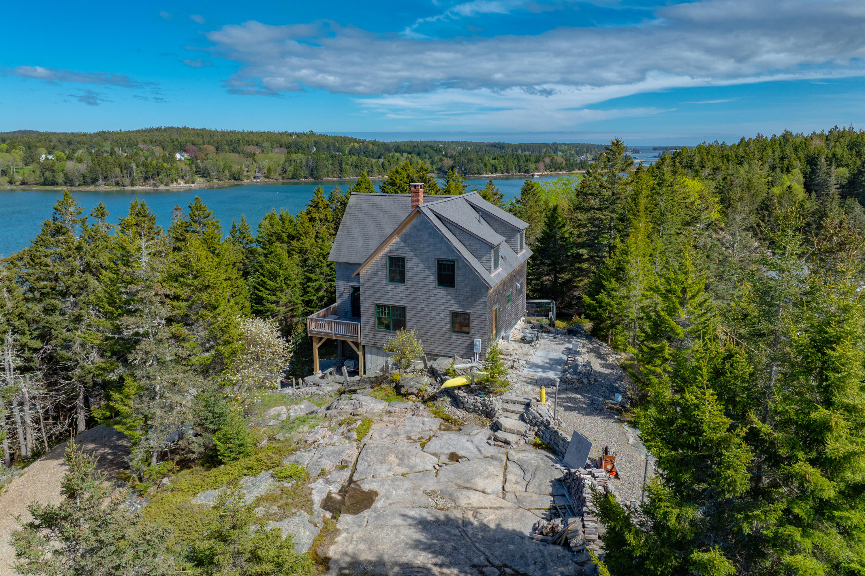 Gray-shingled home on rocky coastal terrain, surrounded by evergreens beside a blue inlet.
