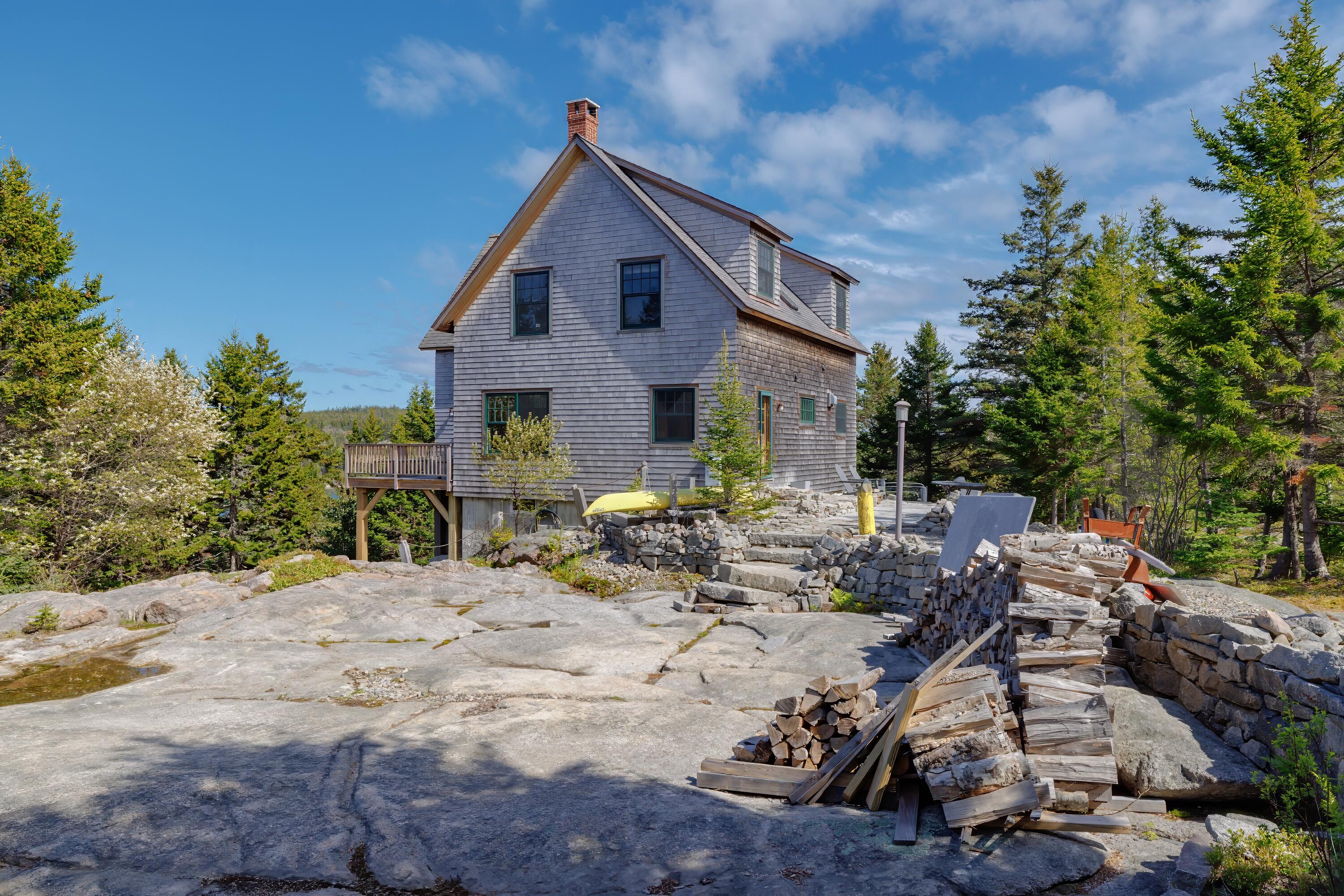 Gray-shingled house with a raised deck on rocky terrain, surrounded by evergreens and stacked firewood.