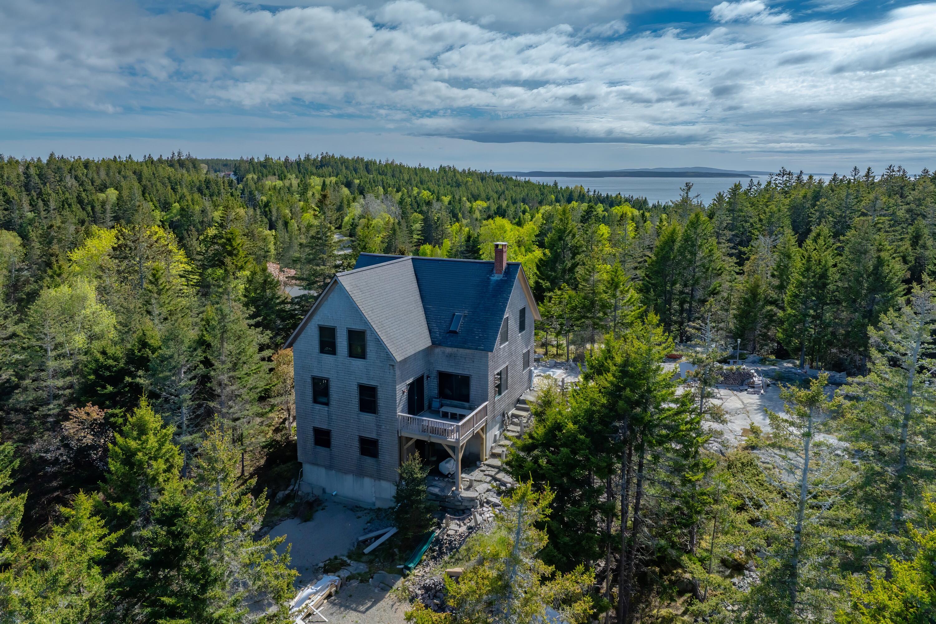 Gray shingle house amid dense woodland, with a deck and distant water view.