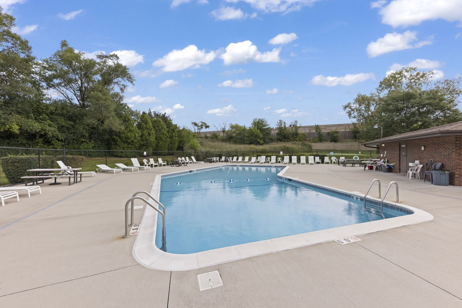 Outdoor swimming pool with a broad concrete deck, lounge chairs, and a brick pool building.