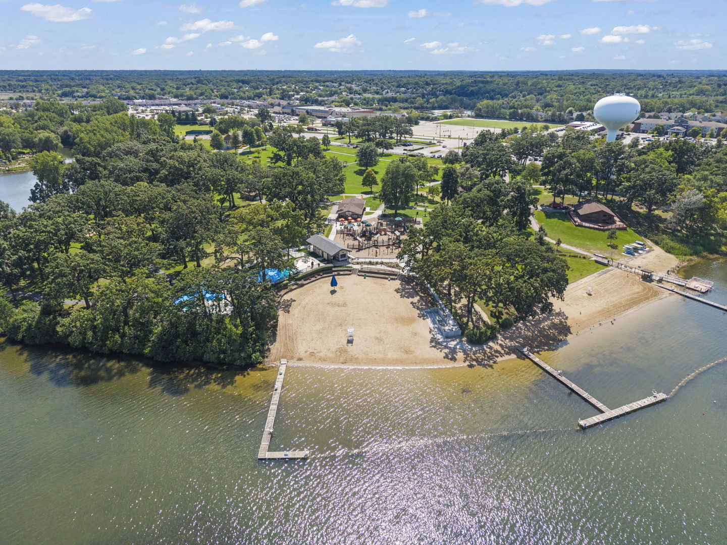 Aerial view of a lakeside beach and recreation area with docks, pool, and wooded grounds.