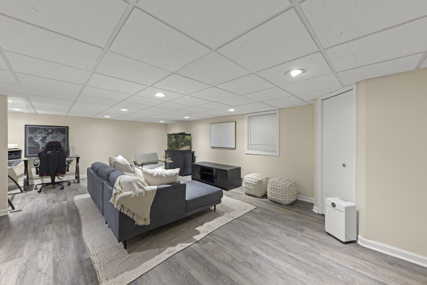 Basement living area with a gray sectional, patterned poufs, and a desk beneath a drop ceiling.