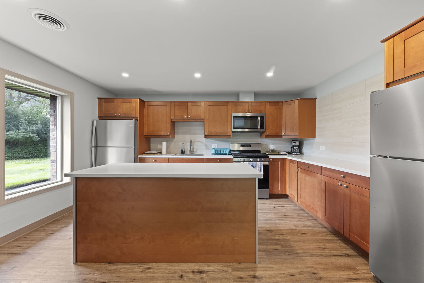 Kitchen with a large island, warm wood cabinetry, stainless steel appliances, and a window overlooking greenery.