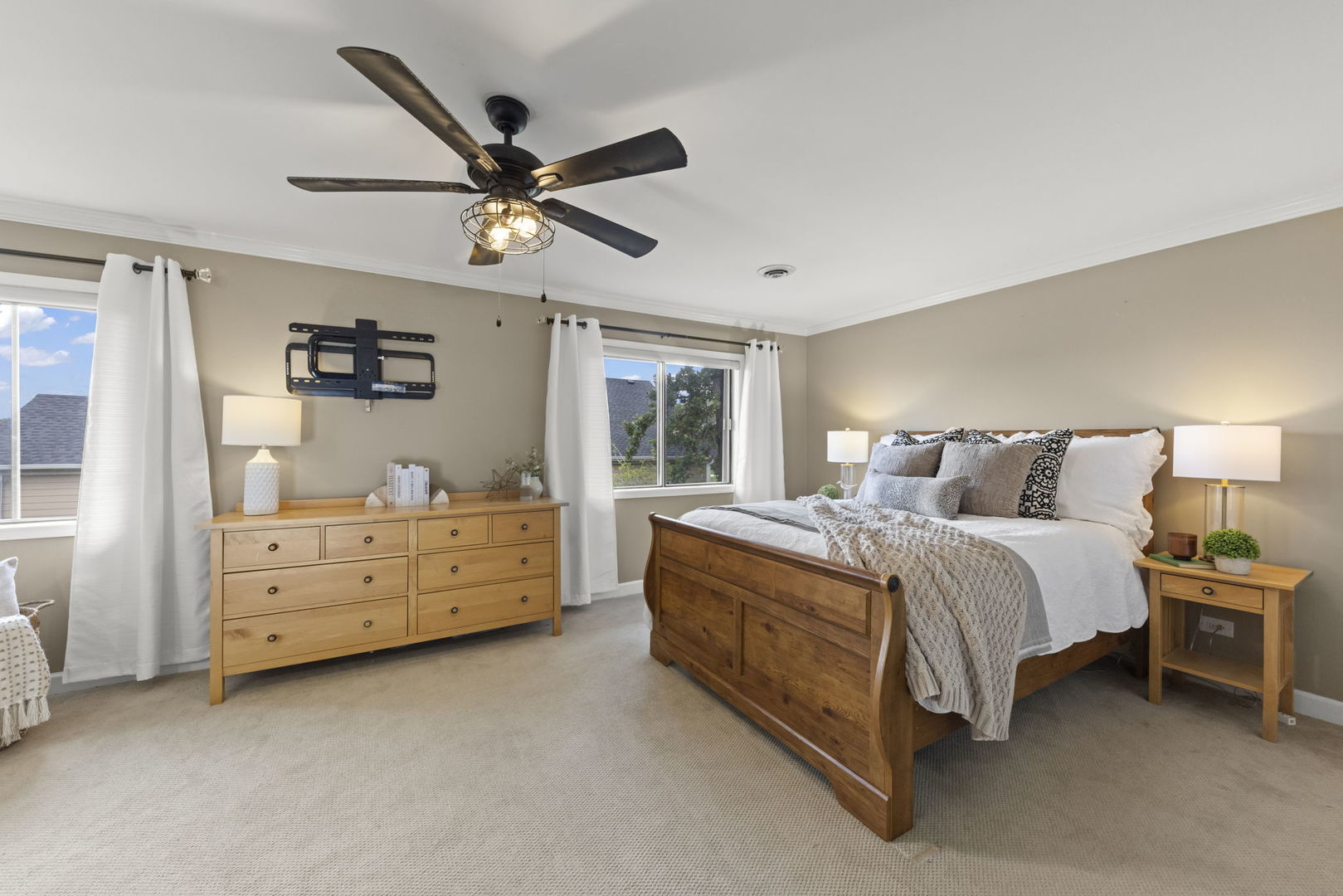 Bedroom with a wooden bed, matching dresser, two curtained windows, and a ceiling fan with a lit cage light.
