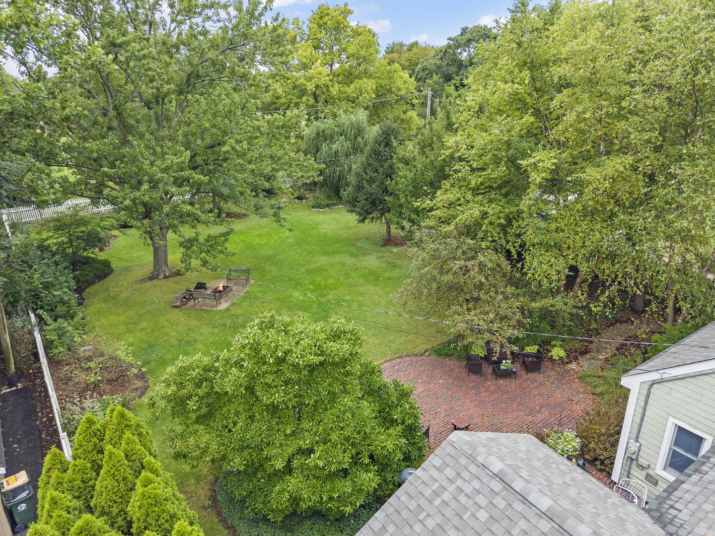 Aerial backyard view with a wide lawn, mature trees, brick patio, outdoor seating, and fire pit.