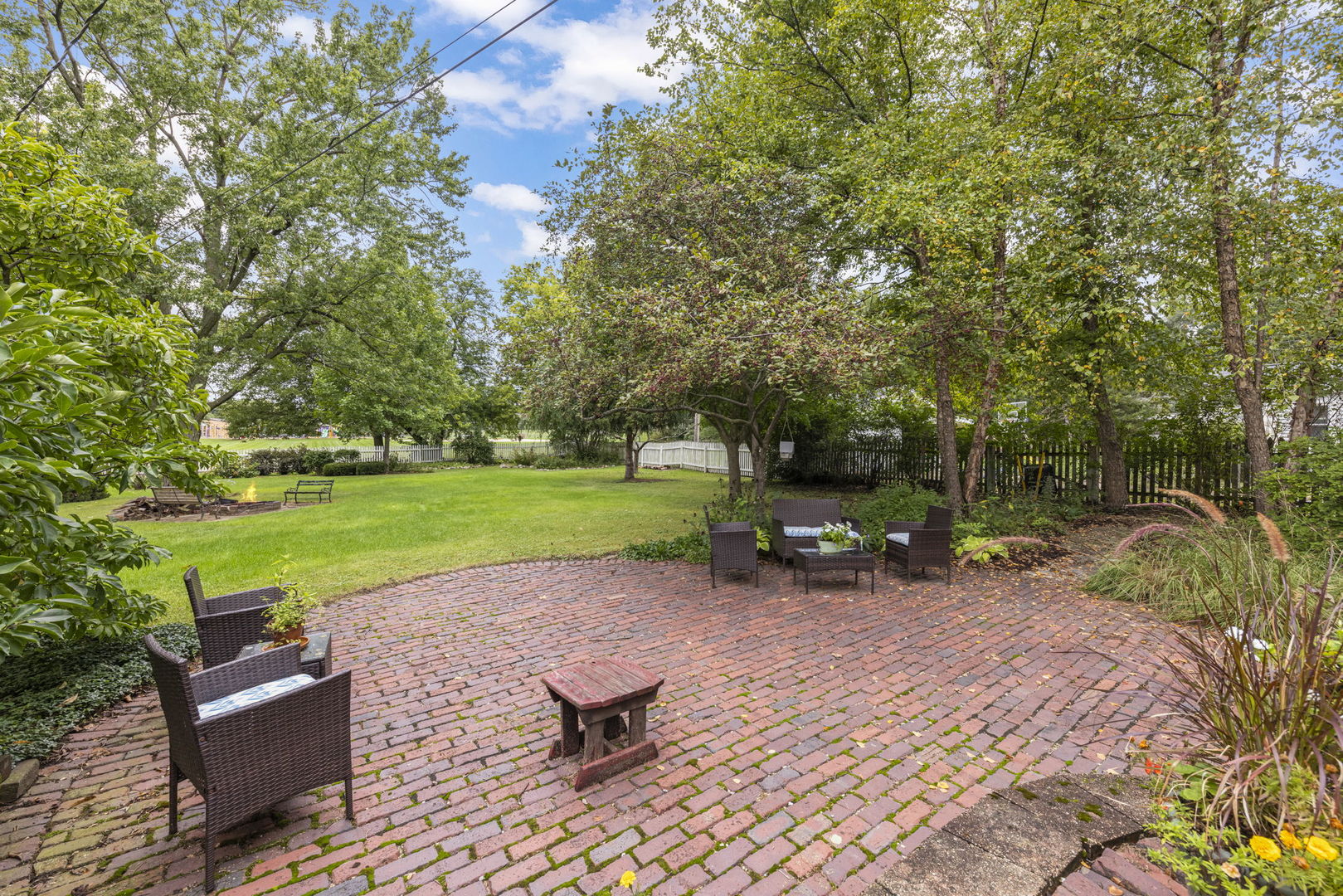 Brick backyard patio with outdoor seating beside a tree-shaded lawn and fire pit.