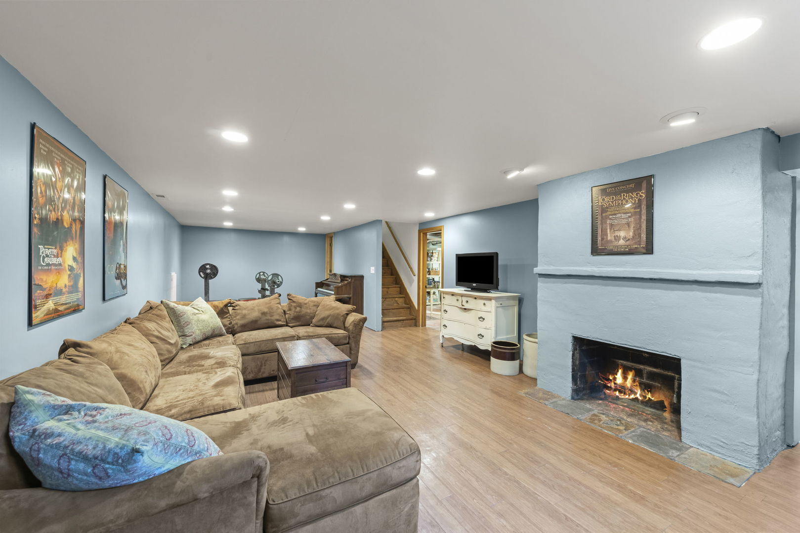Basement family room with a tan sectional, blue walls, stairs, and a lit fireplace.