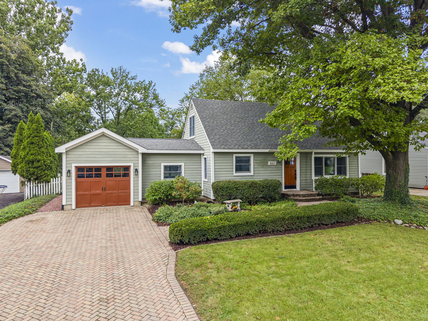 Light gray home with orange-red garage door, brick driveway, landscaped shrubs, and a large shade tree.