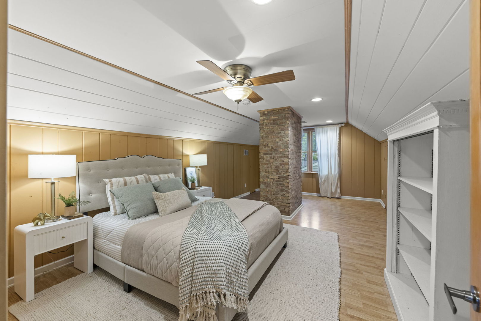 Staged bedroom with a sloped paneled ceiling, exposed brick chimney, and built-in shelves.