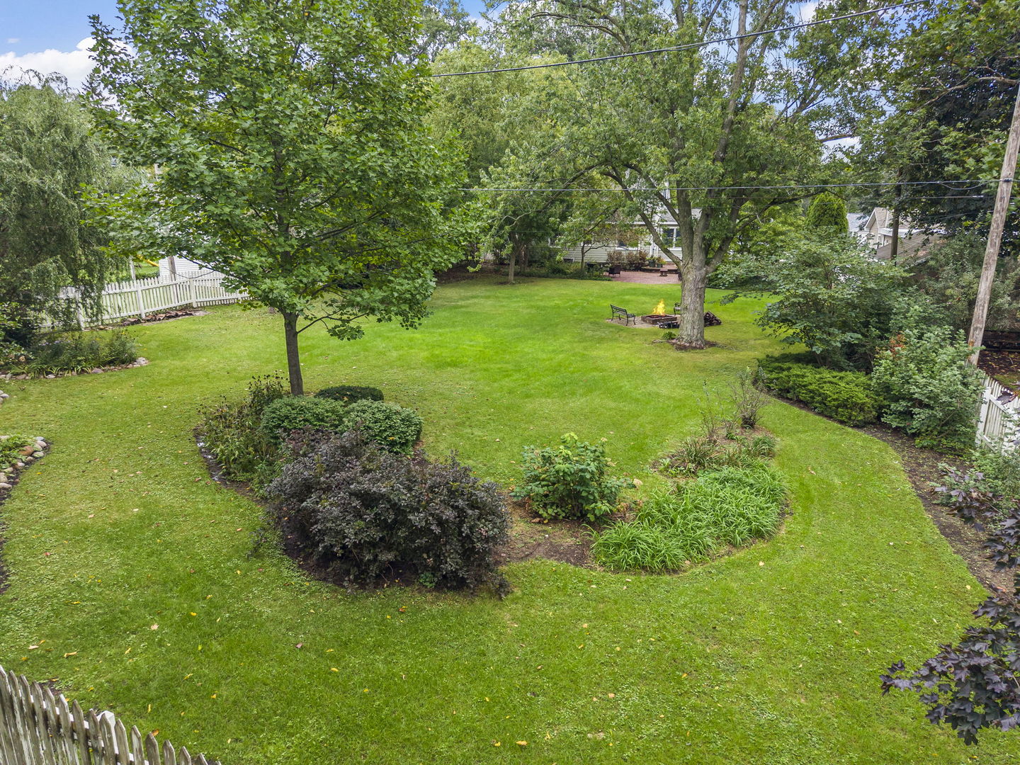 Landscaped backyard with a wide lawn, mature trees, garden beds, and a fire pit seating area.