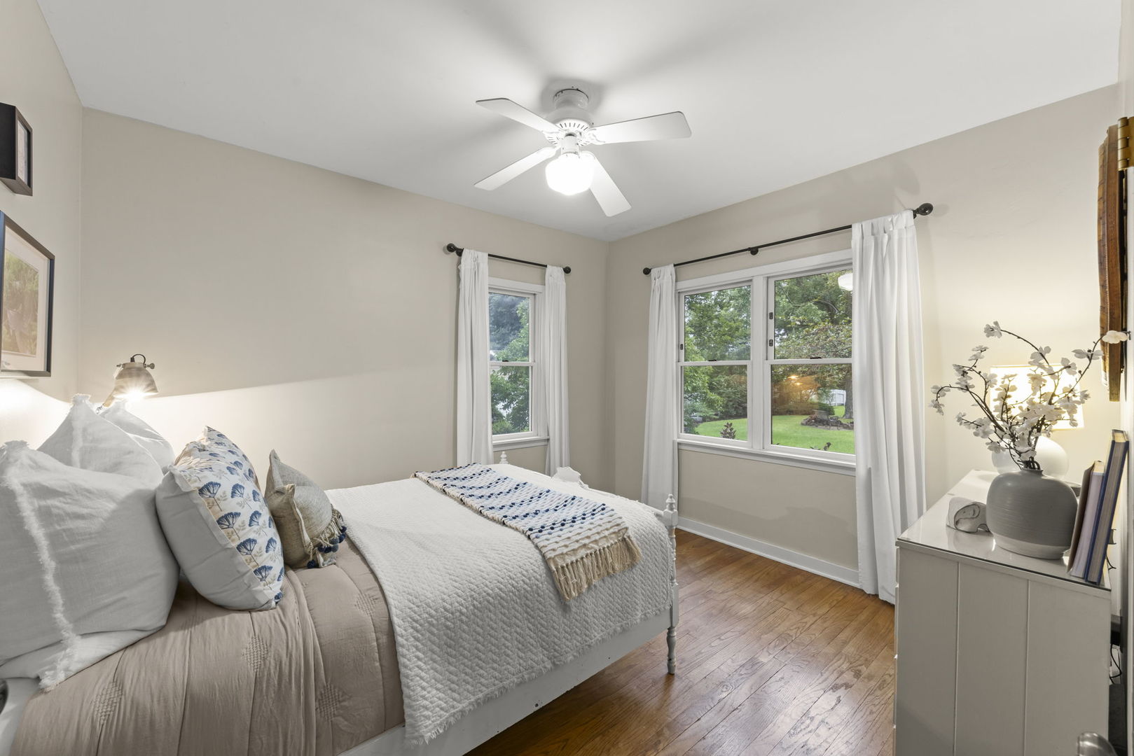 Bedroom with a white-dressed bed, hardwood floors, ceiling fan, and windows overlooking a green yard.