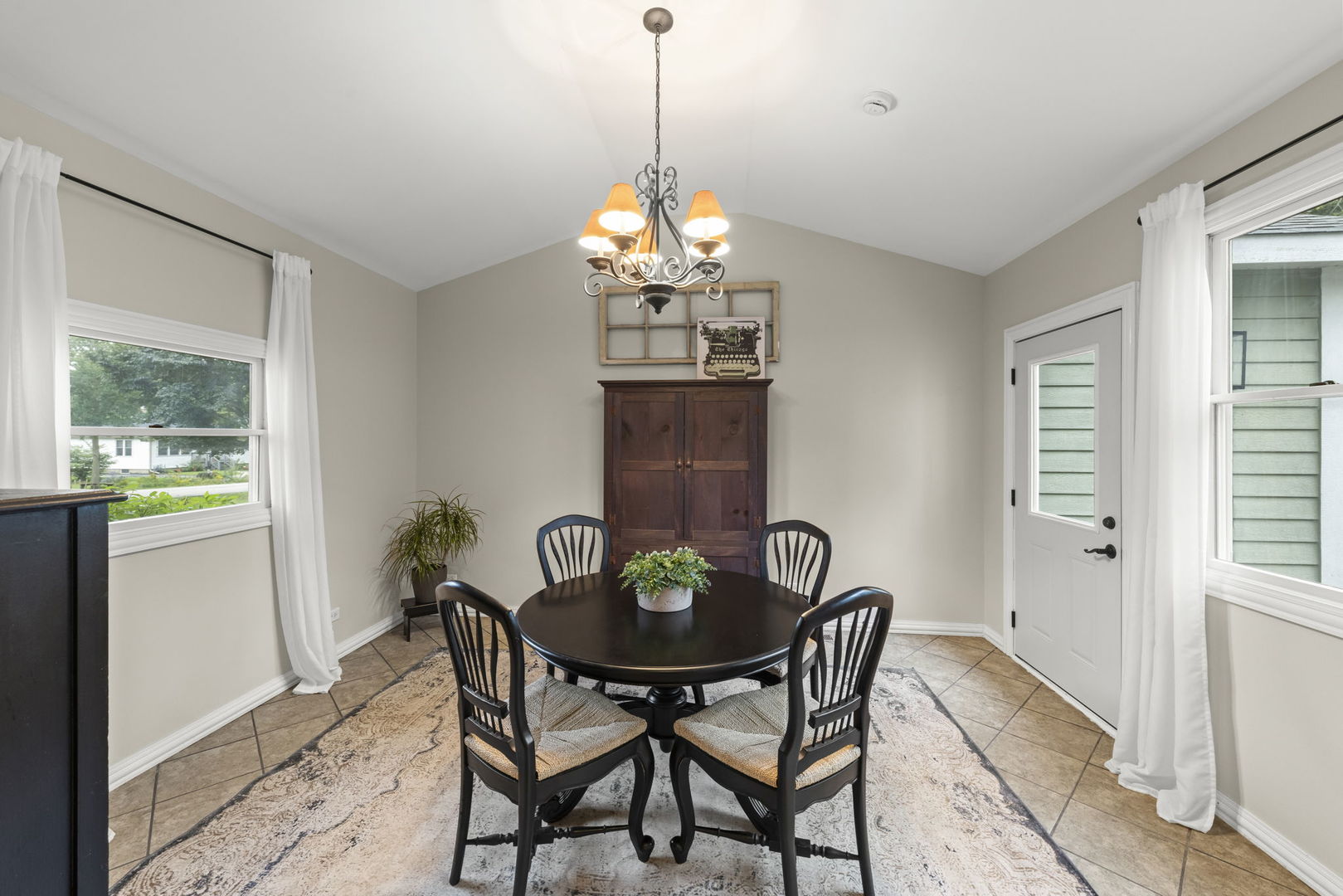 Dining room with a round table and four chairs beneath a chandelier, between two curtained windows.