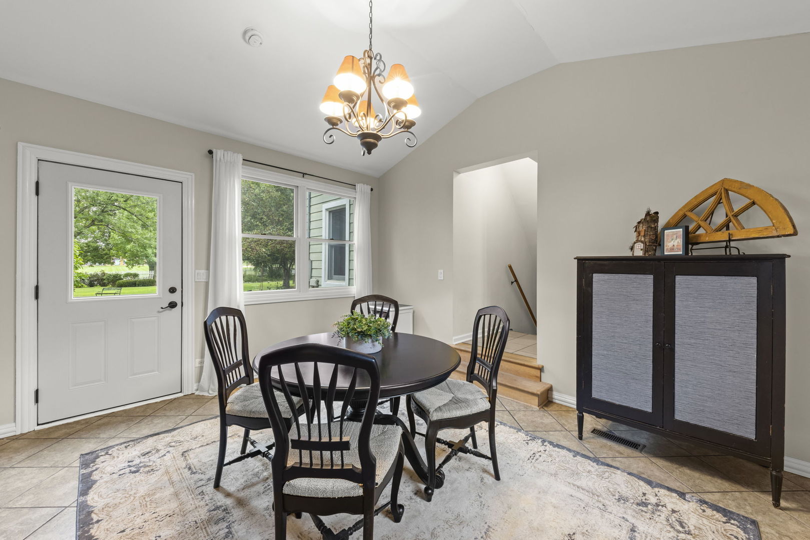 Dining room with a round table, chandelier, tiled floor, and windows overlooking greenery.